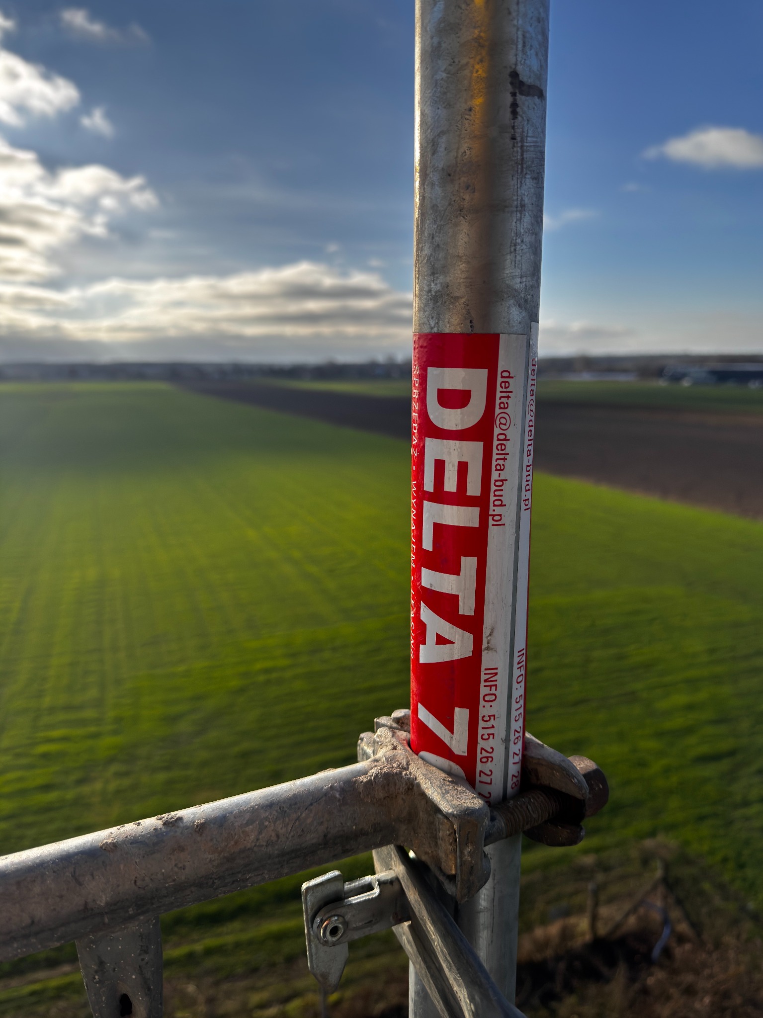 Metalowy słupek rusztowania z naklejką DELTA na tle zielonych pól i błękitnego nieba. Widok z góry, z ostrością na element konstrukcyjny.