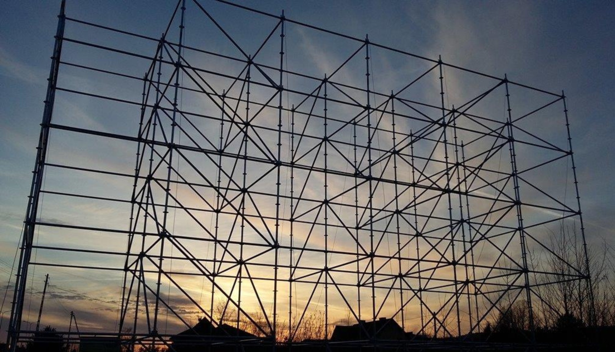 Stalowa konstrukcja pod billboard reklamowy na tle wieczornego nieba. Widoczne elementy zespawane w kratownicę, drzewa i słupy energetyczne w tle, ciepłe światło zachodu.