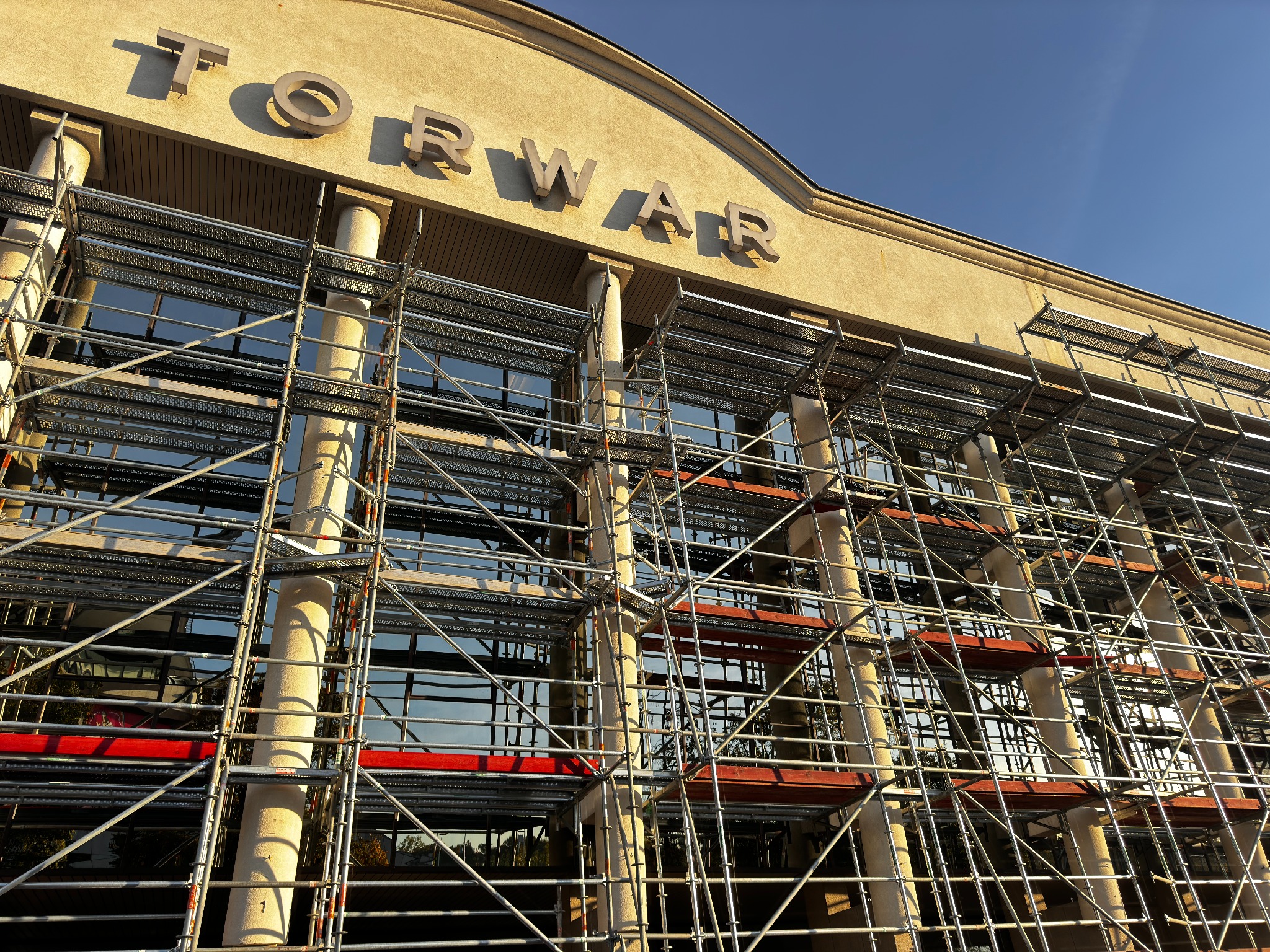 Elewacja budynku Torwar w trakcie prac budowlanych z użyciem metalowych rusztowań. Widok z dołu, błękitne niebo w tle.