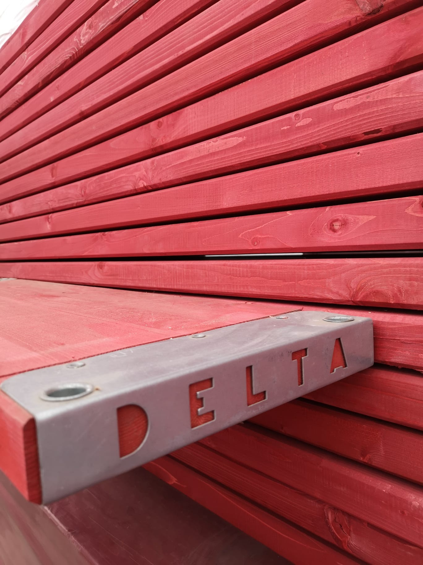 Czerwone, drewniane podesty rusztowaniowe z metalowym wzmocnieniem DELTA, ułożone warstwowo. Widoczne słoje drewna i mocne połączenia.