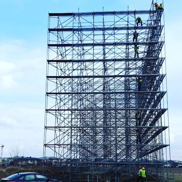 Montaż rusztowania modułowego na tle błękitnego nieba. Na konstrukcji widoczni pracownicy w odblaskowych ubraniach. U dołu czarny samochód osobowy.
