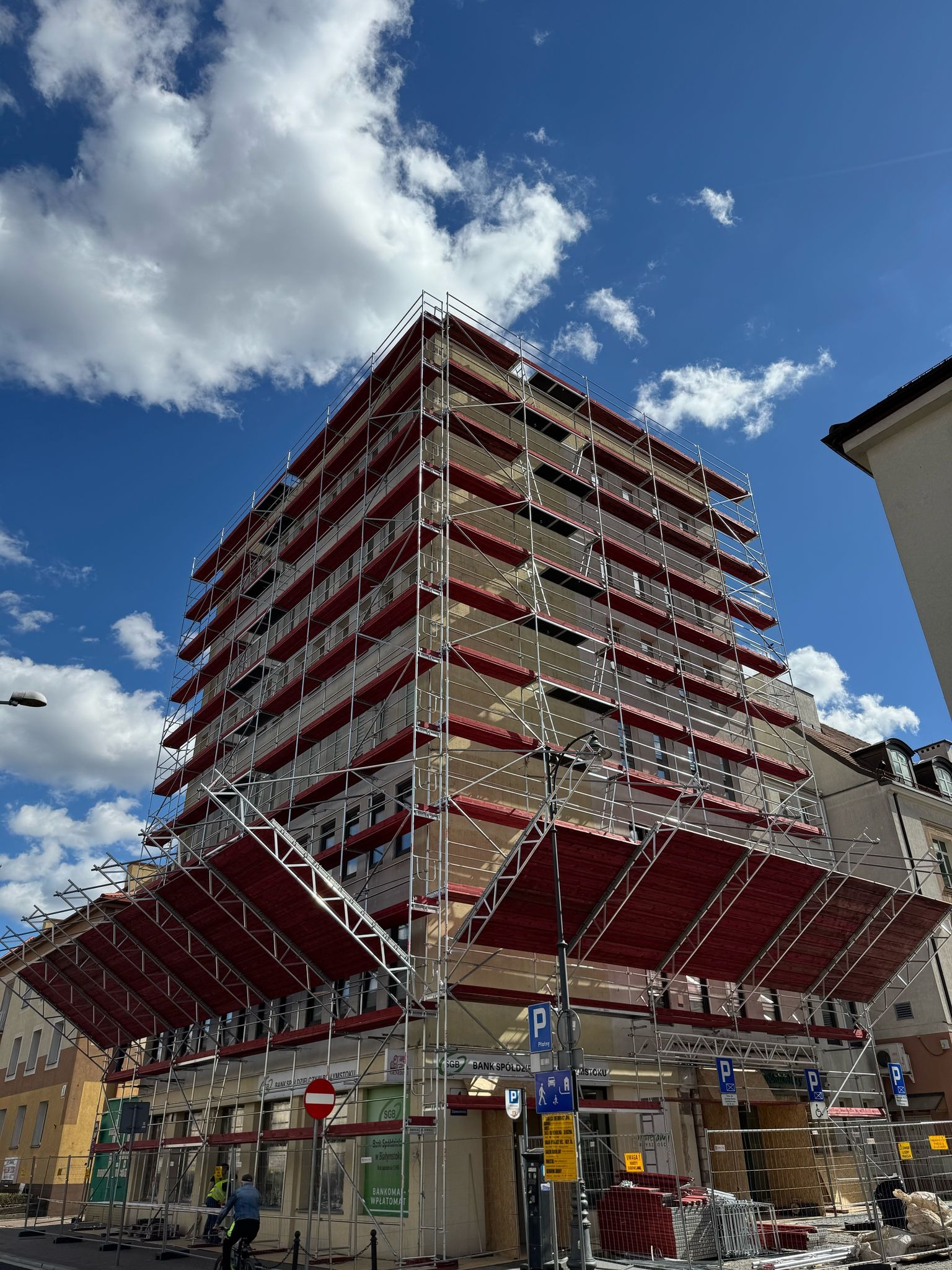 Budynek w rusztowaniach elewacyjnych DELTA, widok z dołu na tle błękitnego nieba z chmurami, Warszawa. Widoczne znaki drogowe i elementy infrastruktury miejskiej.