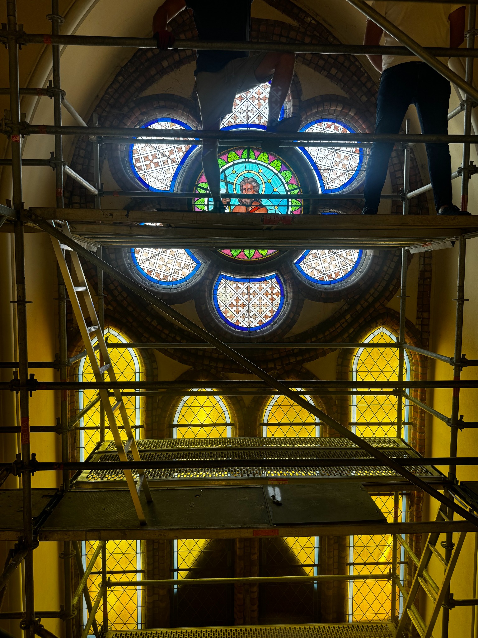 Rusztowanie DELTA wewnątrz budynku z witrażami. Widok z dołu na konstrukcję i fragmenty okien. Prace konserwatorskie w toku, elementy rusztowania z drabinką.
