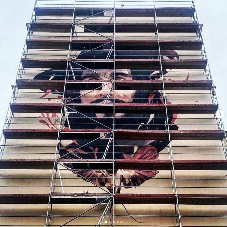 Rusztowanie modułowe na tle muralu z portretem mężczyzny, widok z dołu. Elementy rusztowania tworzą siatkę na ścianie budynku.