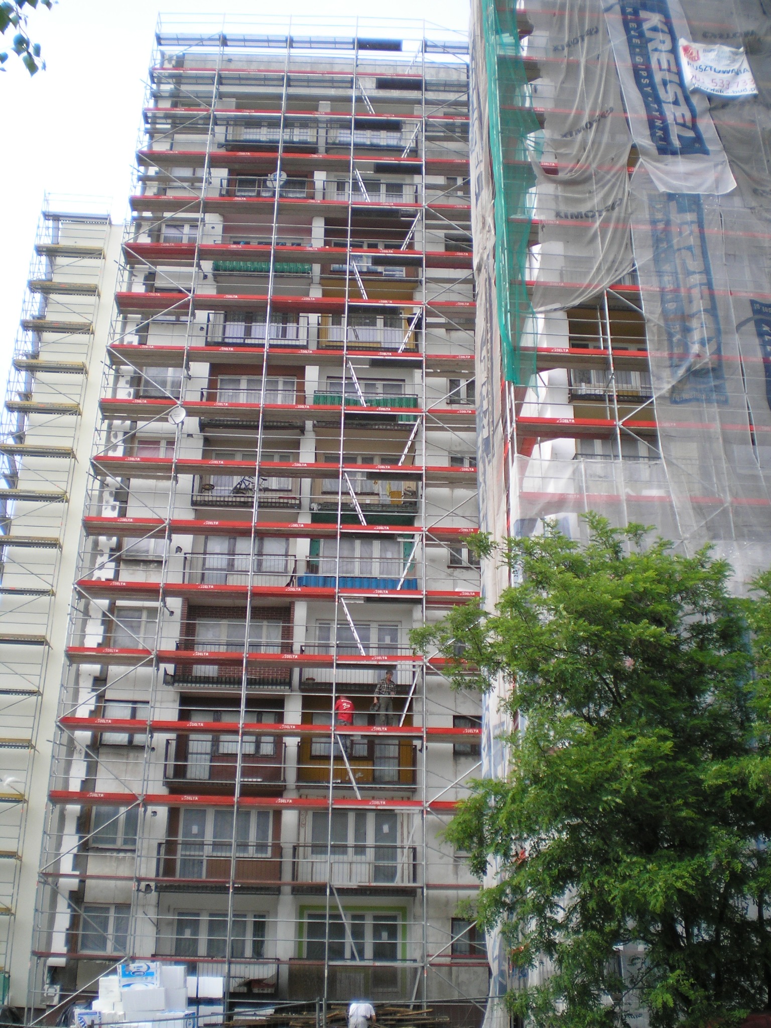 Wielopiętrowy budynek w rusztowaniach z czerwonymi poprzeczkami, częściowo osłonięty siatką ochronną, widok z dołu, fragment drzewa po prawej.