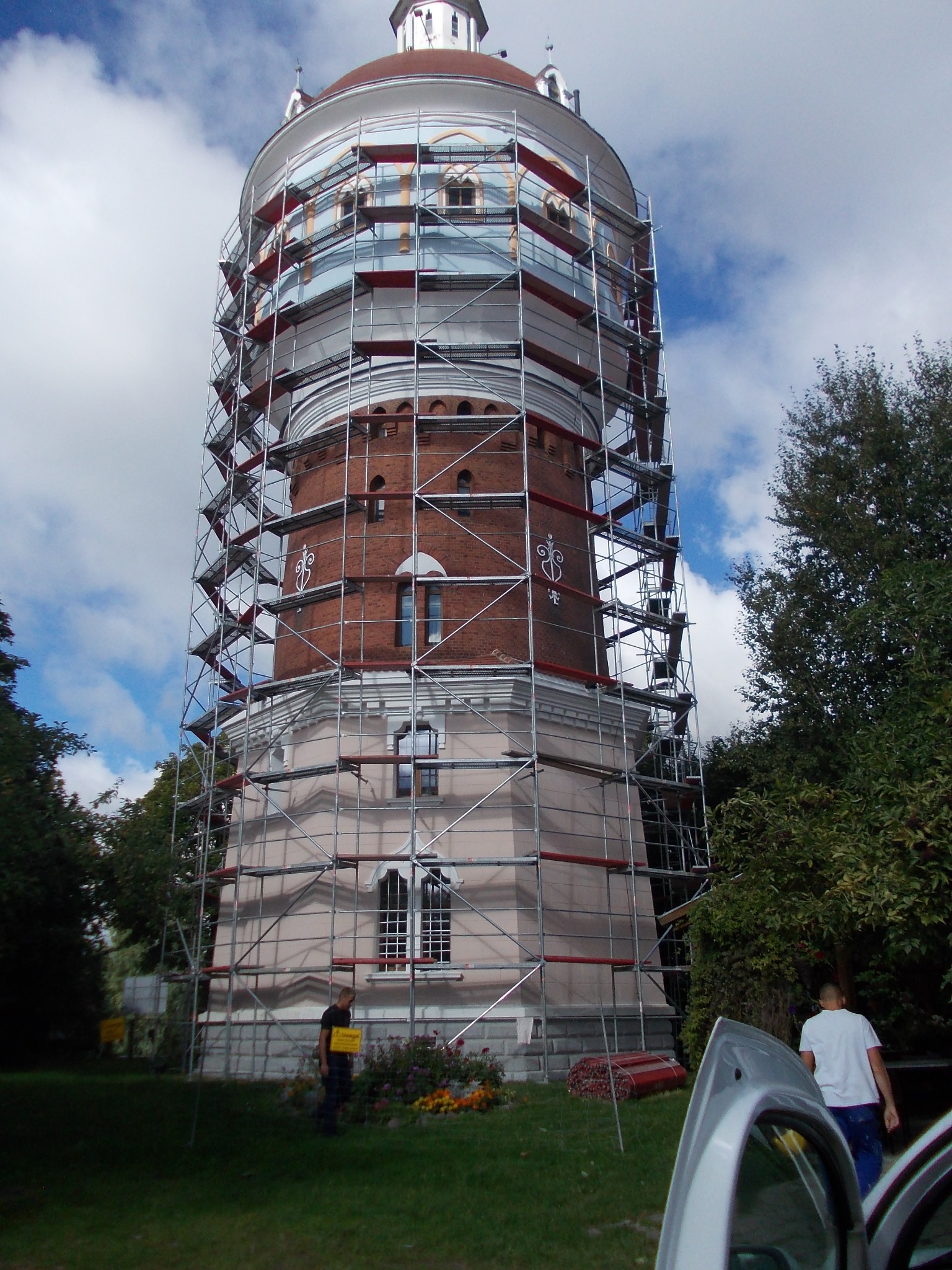 Wieża ciśnień w rusztowaniach, widoczni dwaj mężczyźni. Budowla z cegły i tynku, na tle nieba z chmurami. Elementy rusztowania w kolorze srebrnym i czerwonym.