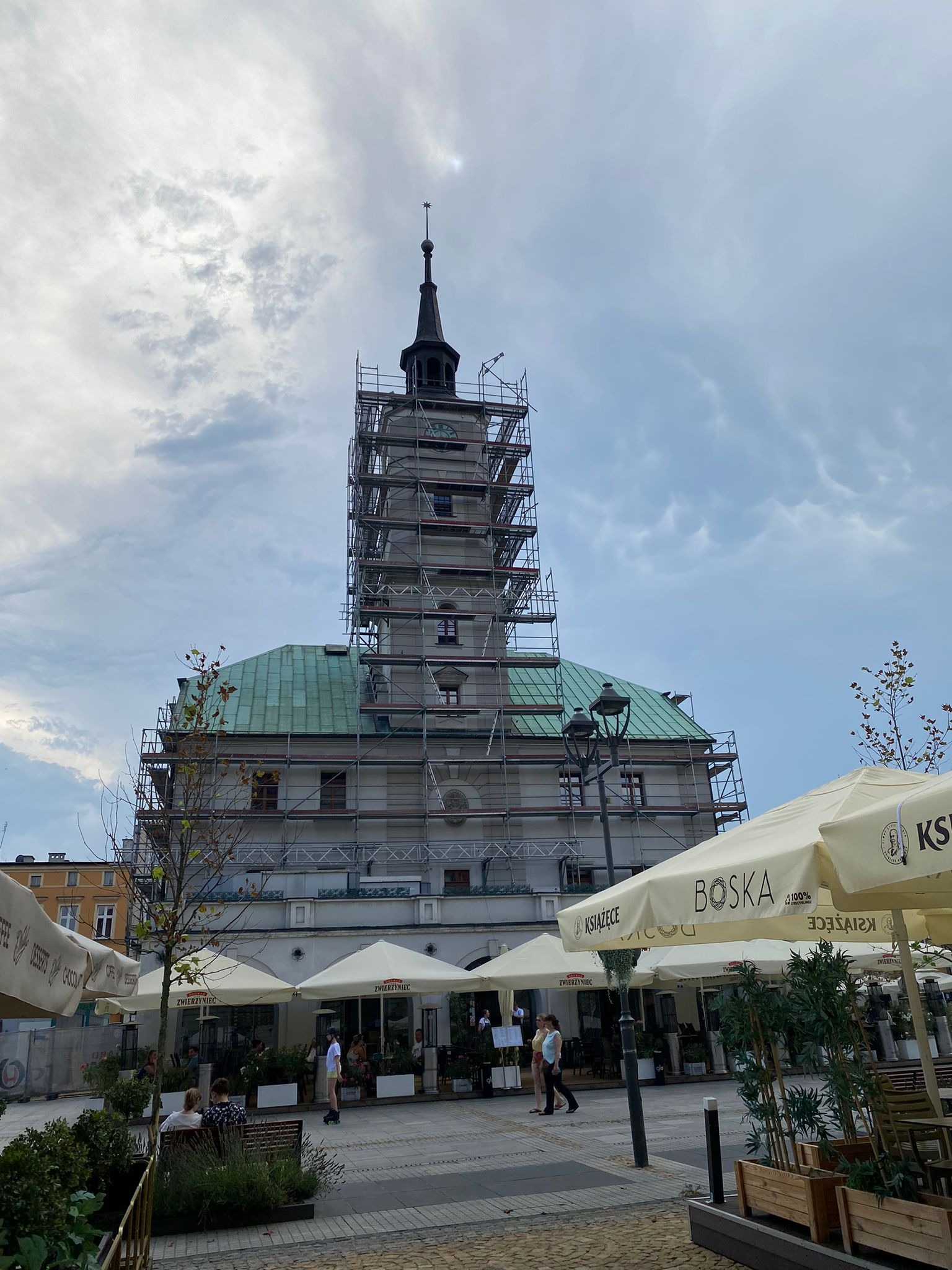 Ratusz w rusztowaniach, widok z placu miejskiego z parasolami kawiarnianymi w słoneczny dzień. Prace remontowe w toku.