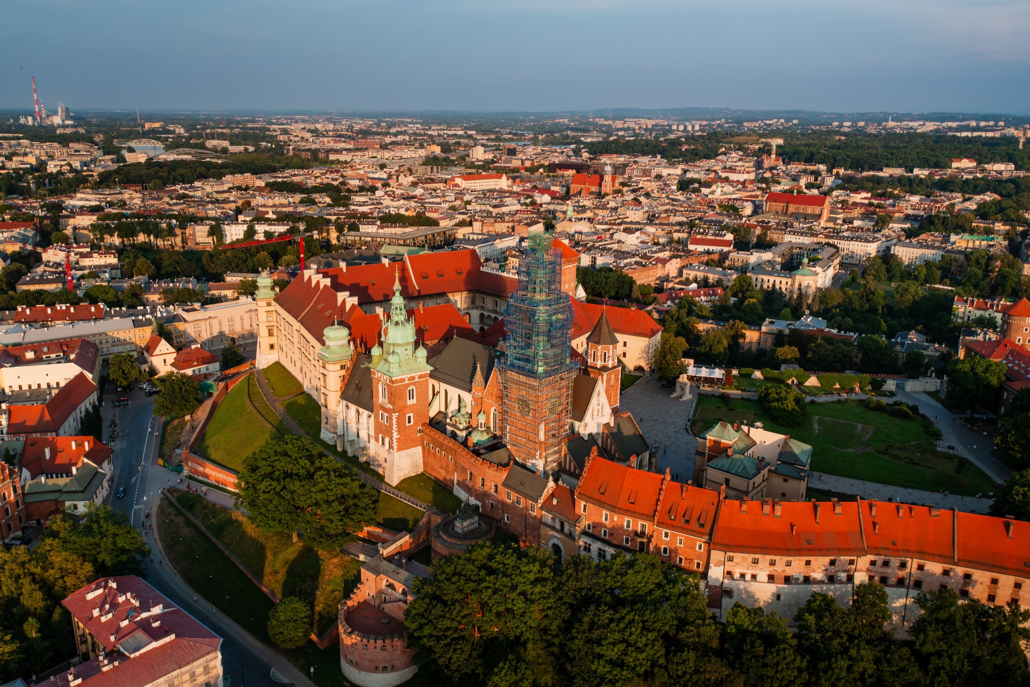 Kraków z lotu ptaka: Wawel w rusztowaniach na tle panoramy miasta, ciepłe światło zachodzącego słońca.