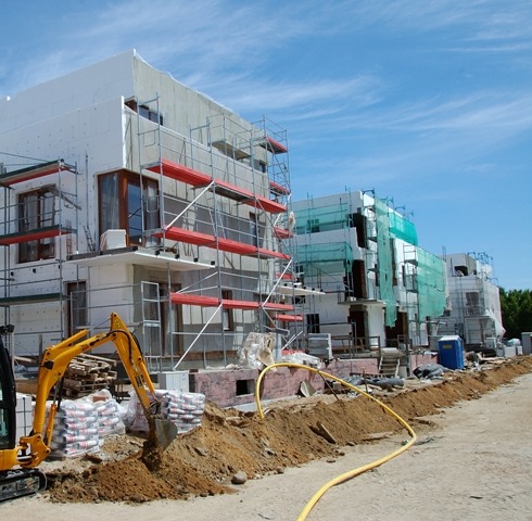 Budowa osiedla domów jednorodzinnych w trakcie ocieplania, z rusztowaniami i minikoparką. Widok na budynki w jasnych kolorach pod błękitnym niebem.