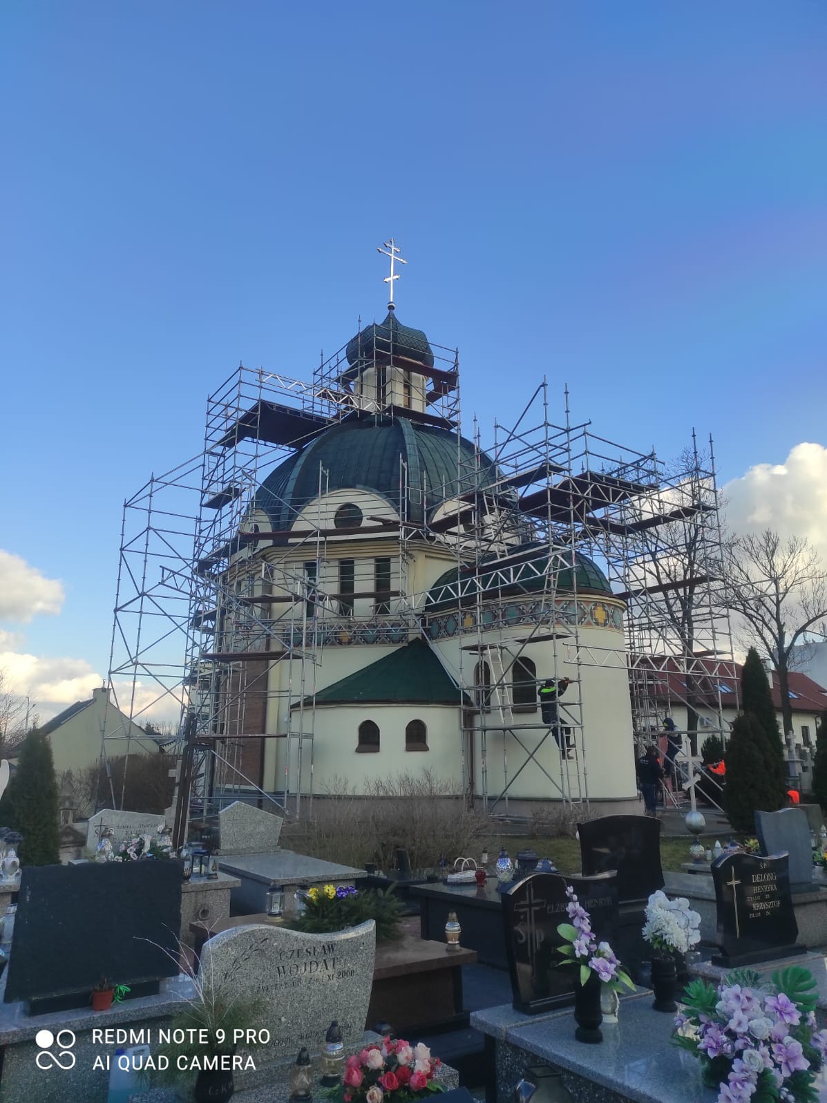 Kościół z zieloną kopułą w rusztowaniach, widok z dołu, cmentarz na pierwszym planie. Prace konserwatorskie na zabytkowym obiekcie sakralnym.