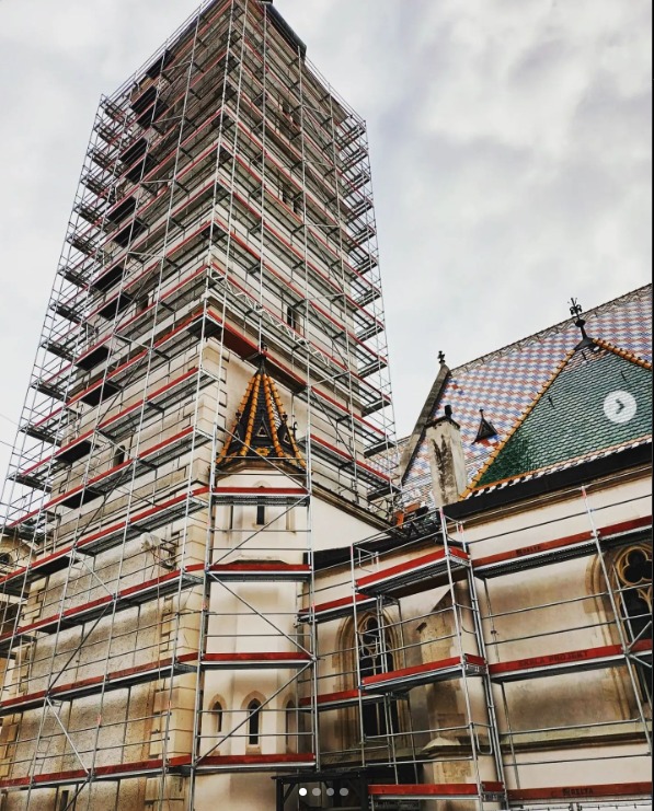 Zabytkowy kościół w rusztowaniach, widok z dołu. Rusztowania specjalistyczne na historycznej elewacji, częściowo zasłaniają charakterystyczny dach z wzorem.