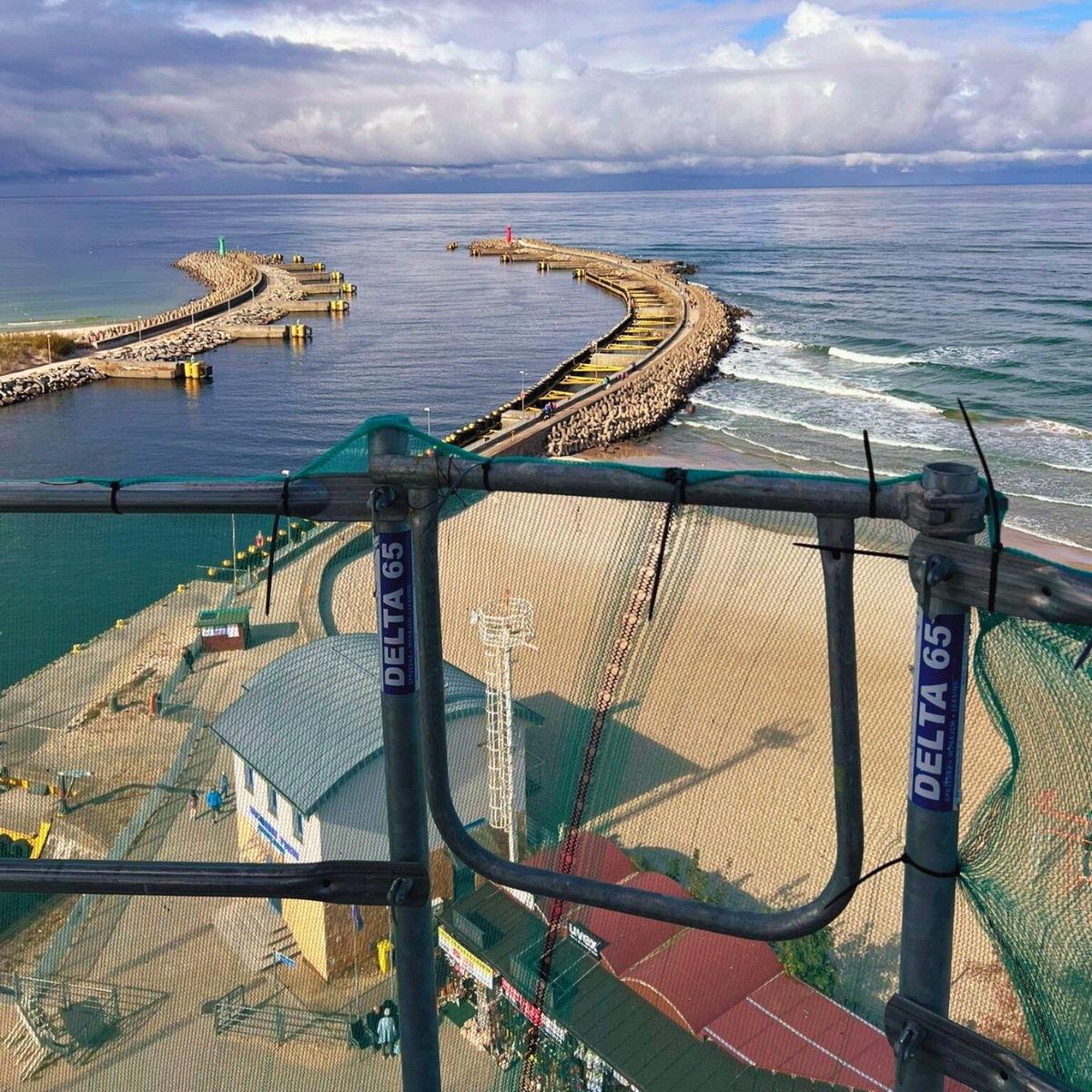 Widok z rusztowania Delta 65 na port z falochronami, morze i plażę. Perspektywa z góry, pochmurne niebo. Rusztowanie z siatką ochronną na pierwszym planie.