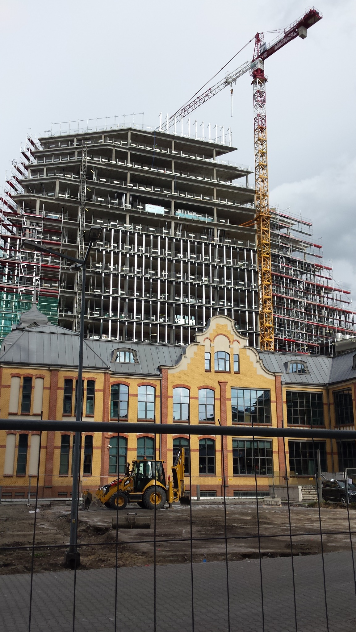 Żółta koparko-ładowarka na placu budowy przed starym budynkiem i konstrukcją wieżowca w rusztowaniach, widok przez metalowe ogrodzenie.