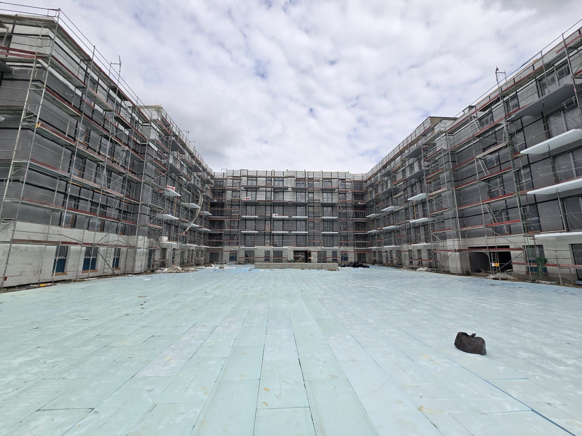 Budynek w trakcie budowy, otoczony rusztowaniami, z widoczną izolacją podłogową i niebem w tle. Perspektywa z poziomu podłoża.