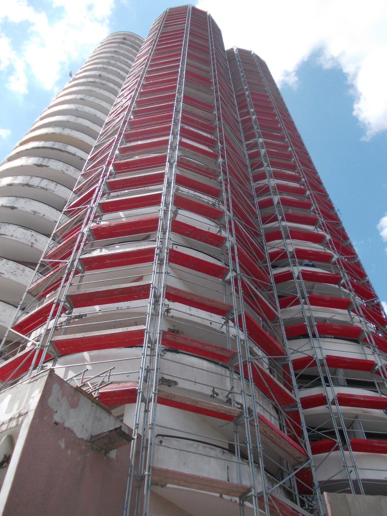 Wysoki budynek w rusztowaniach elewacyjnych z czerwonymi elementami, widziany z dołu pod kątem, na tle błękitnego nieba z chmurami.