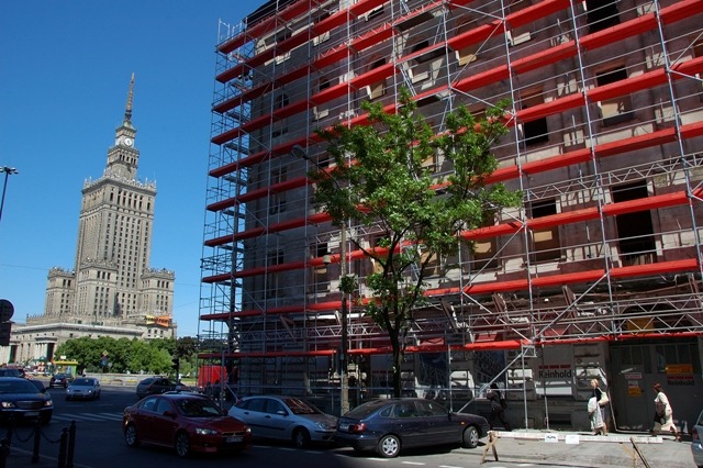 Warszawa: budynek w rusztowaniach DELTA na tle Pałacu Kultury, widok z ulicy, auta, drzewo. Czerwone poprzeczki rusztowań.