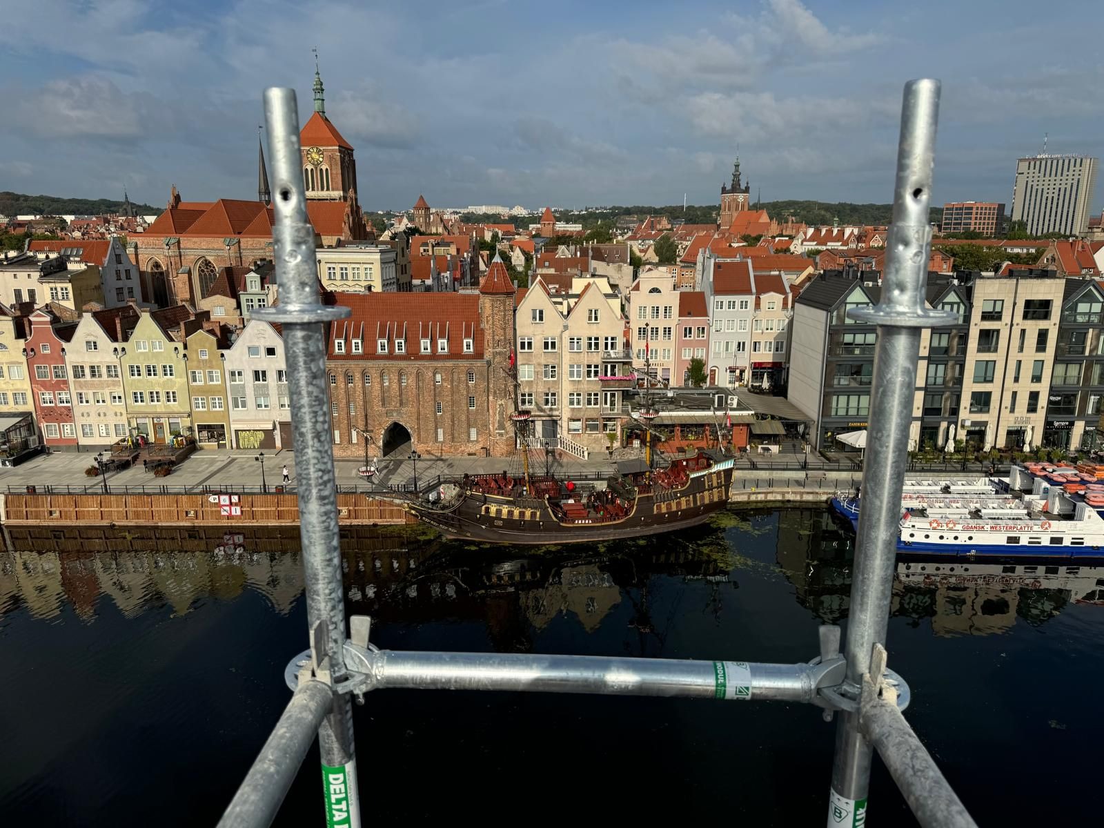 Widok na Gdańsk z rusztowania elewacyjnego, panorama miasta z rzeką, statek stylizowany na galeon, ujęcie z góry przez elementy konstrukcji rusztowania.