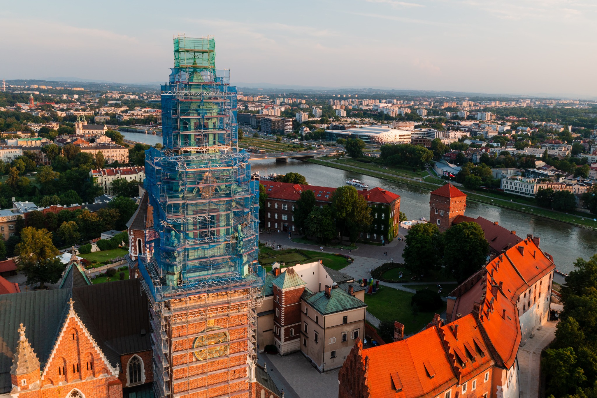 Kraków: Wawel w rusztowaniach, widok z góry na zabytek w trakcie prac konserwatorskich, rzeka Wisła i panorama miasta w tle, ciepłe światło zachodzącego słońca.