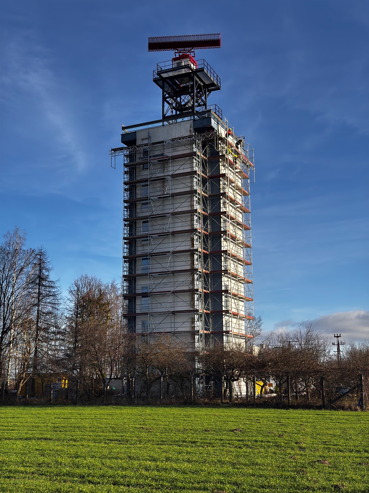 Wysoka wieża w rusztowaniach na tle błękitnego nieba, otoczona zielonym polem i drzewami. Elementy konstrukcyjne na szczycie. Prace budowlane w toku.