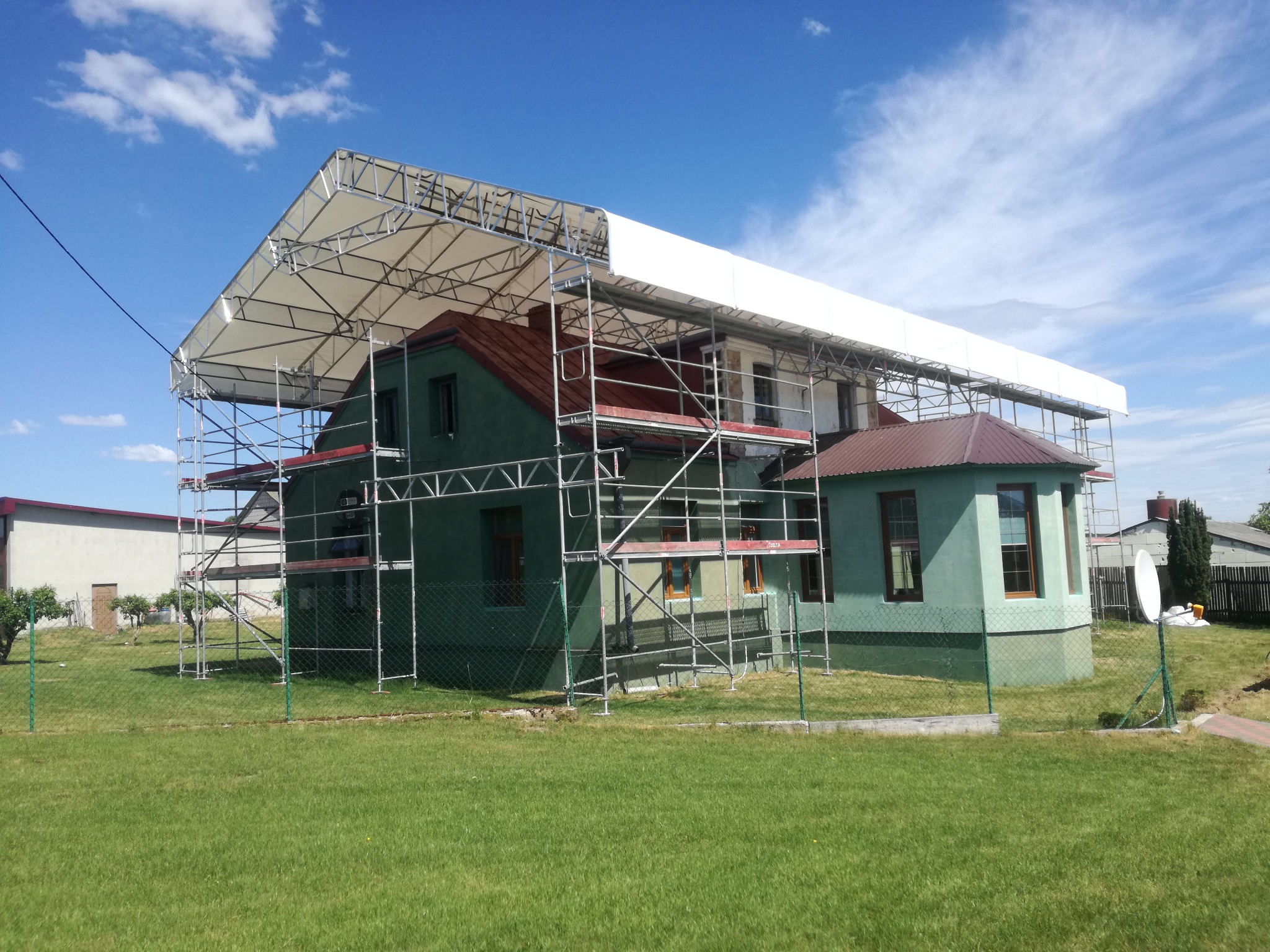 Dom w trakcie remontu z zadaszeniem tymczasowym na rusztowaniu DELTA, widoczny trawnik i niebieskie niebo. Zabezpieczenie konstrukcji przed deszczem lub śniegiem.