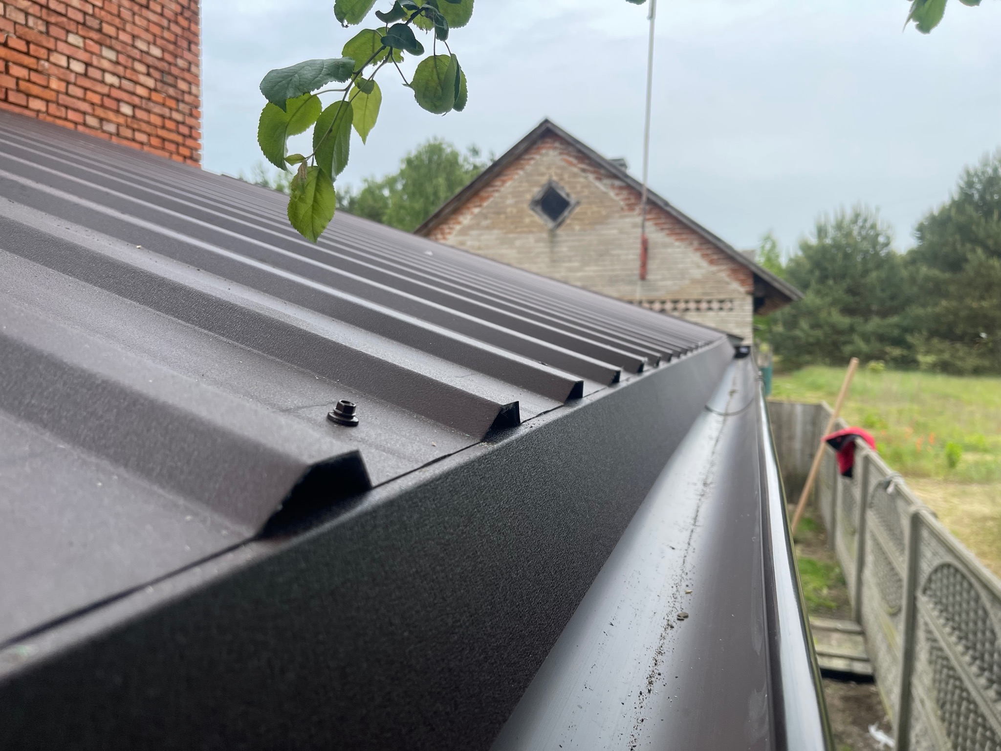 Nowe, brązowe krycie dachu z blachy trapezowej i rynna w Ożarowie. Widoczny komin i fragment sąsiedniego budynku na tle zieleni.