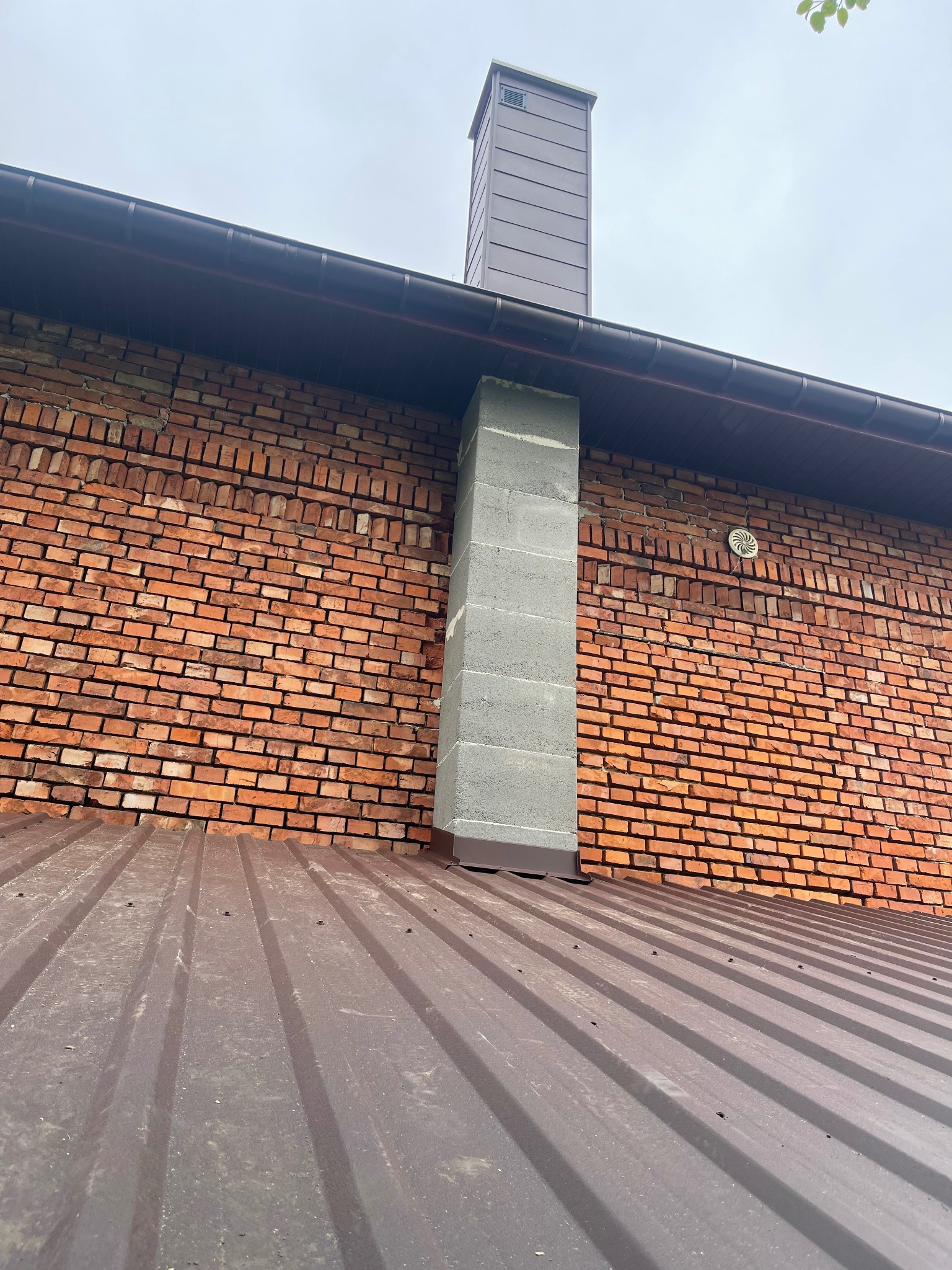 Brązowy dach z blachy trapezowej, szary komin z cegieł i pustaków na tle ceglanej ściany budynku z brązową rynną. Widok z dołu, pochmurne niebo.