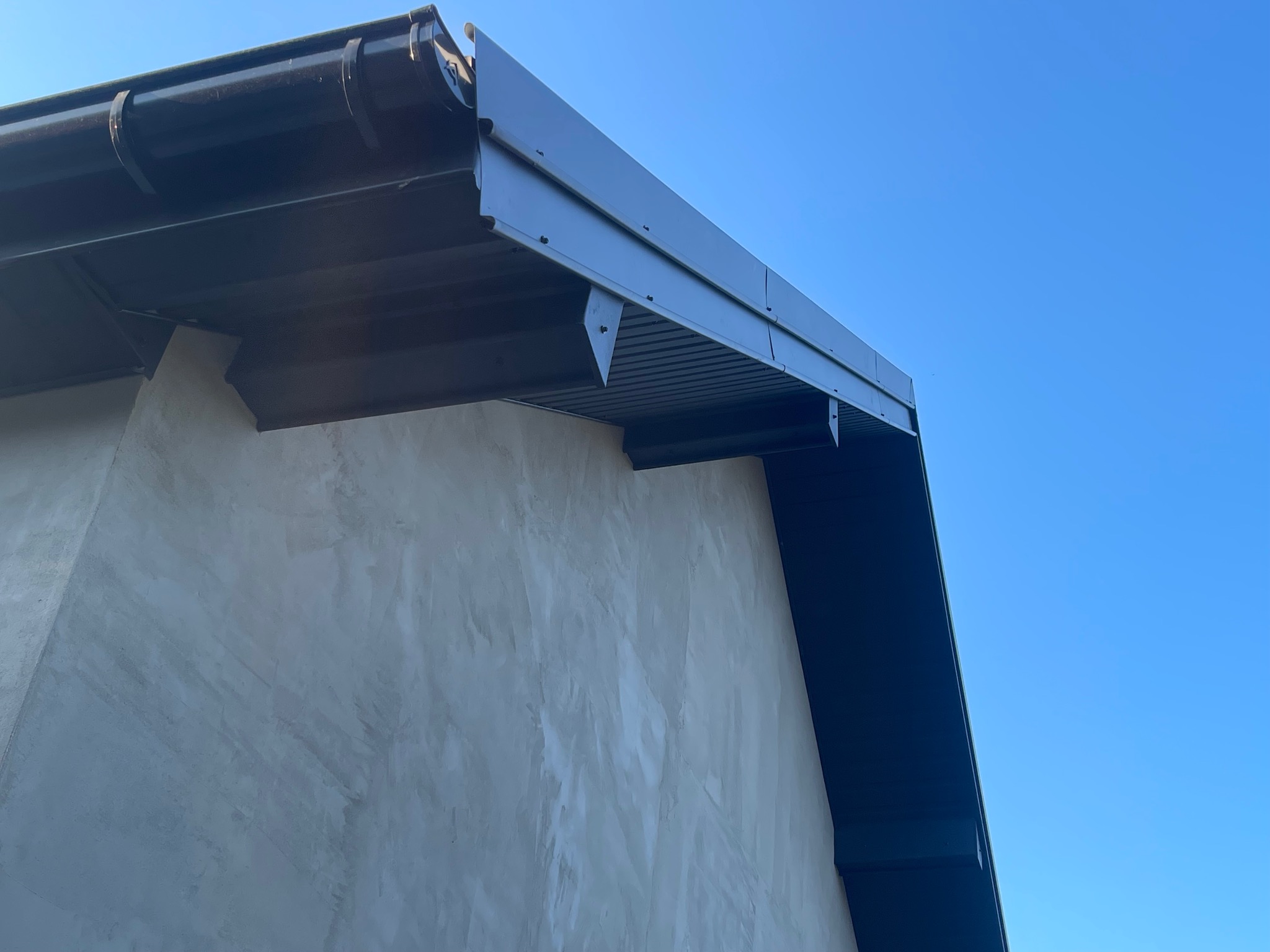Fragment elewacji budynku z zamontowanym, ciemnym orynnowaniem i podbitką dachową, sfotografowany pod kątem, na tle błękitnego nieba.