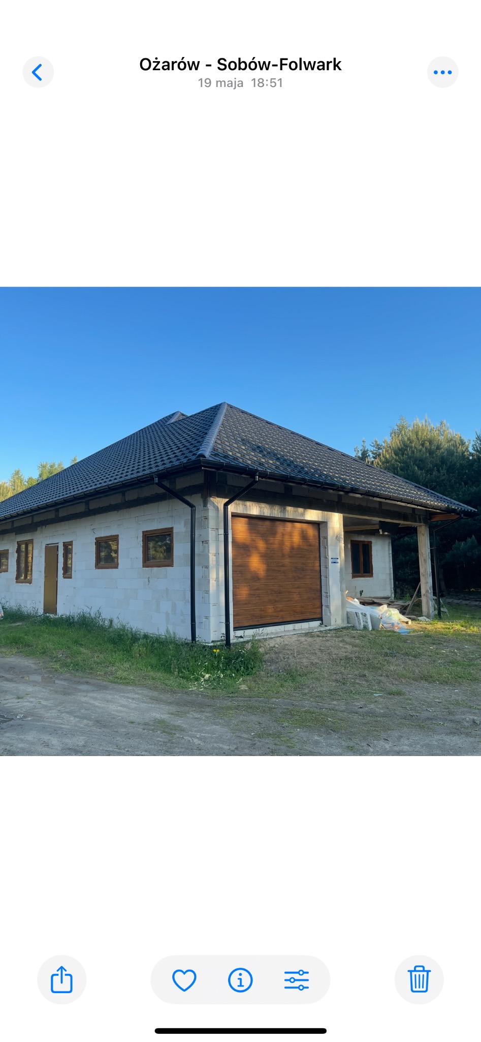 Niedokończony budynek z dachem z blachy na tle zieleni, widoczne okna i brama garażowa. Budynek w stanie surowym, ściany z bloczków.