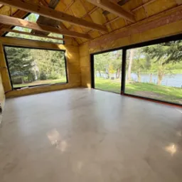 Jasnoszara posadzka z mikrocementu w surowym wnętrzu z oknami na jezioro. Widoczne ocieplenie ścian i drewniana konstrukcja dachu.