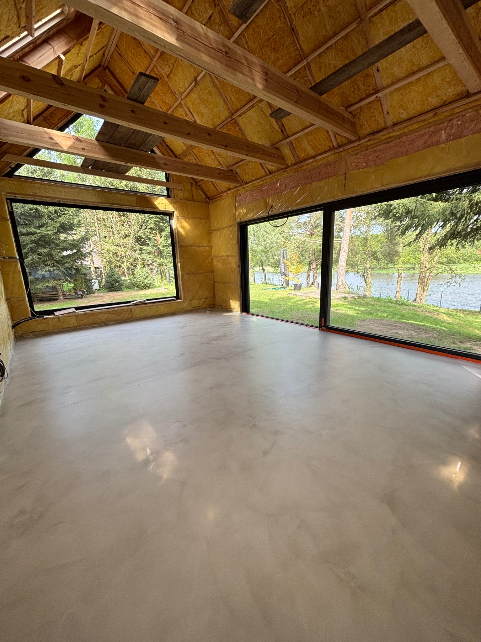 Jasnoszara posadzka z mikrocementu w surowym wnętrzu z oknami na jezioro. Widoczne ocieplenie ścian i drewniana konstrukcja dachu.