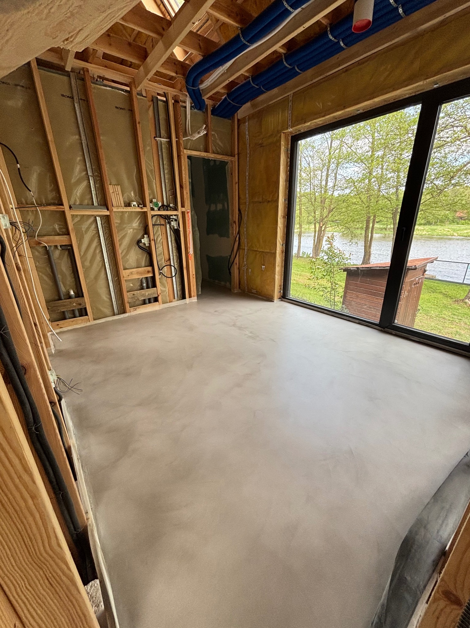 Wnętrze w trakcie wykańczania: gładka, beżowa posadzka, surowe ściany z izolacją, duże okno z widokiem na zieleń i wodę. Niebieskie rury pod sufitem.