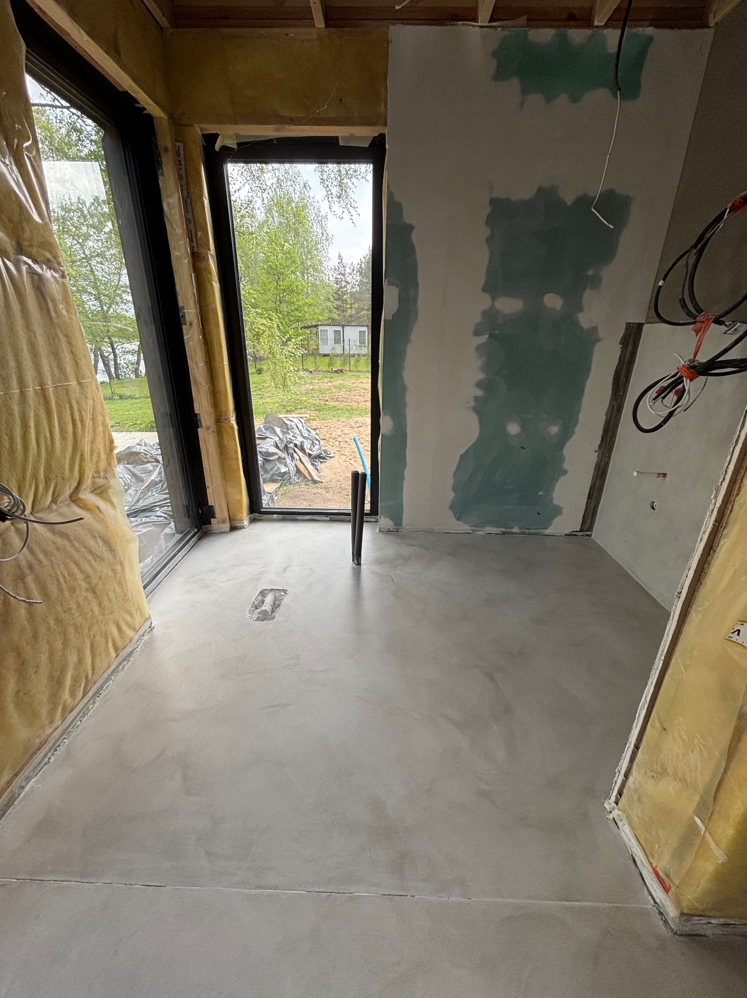 Wnętrze w trakcie wykańczania z szarą, gładką posadzką. Widoczne okna z widokiem na zieleń, ściany przygotowane do malowania i izolacja. Na podłodze ślady po narzędziach.