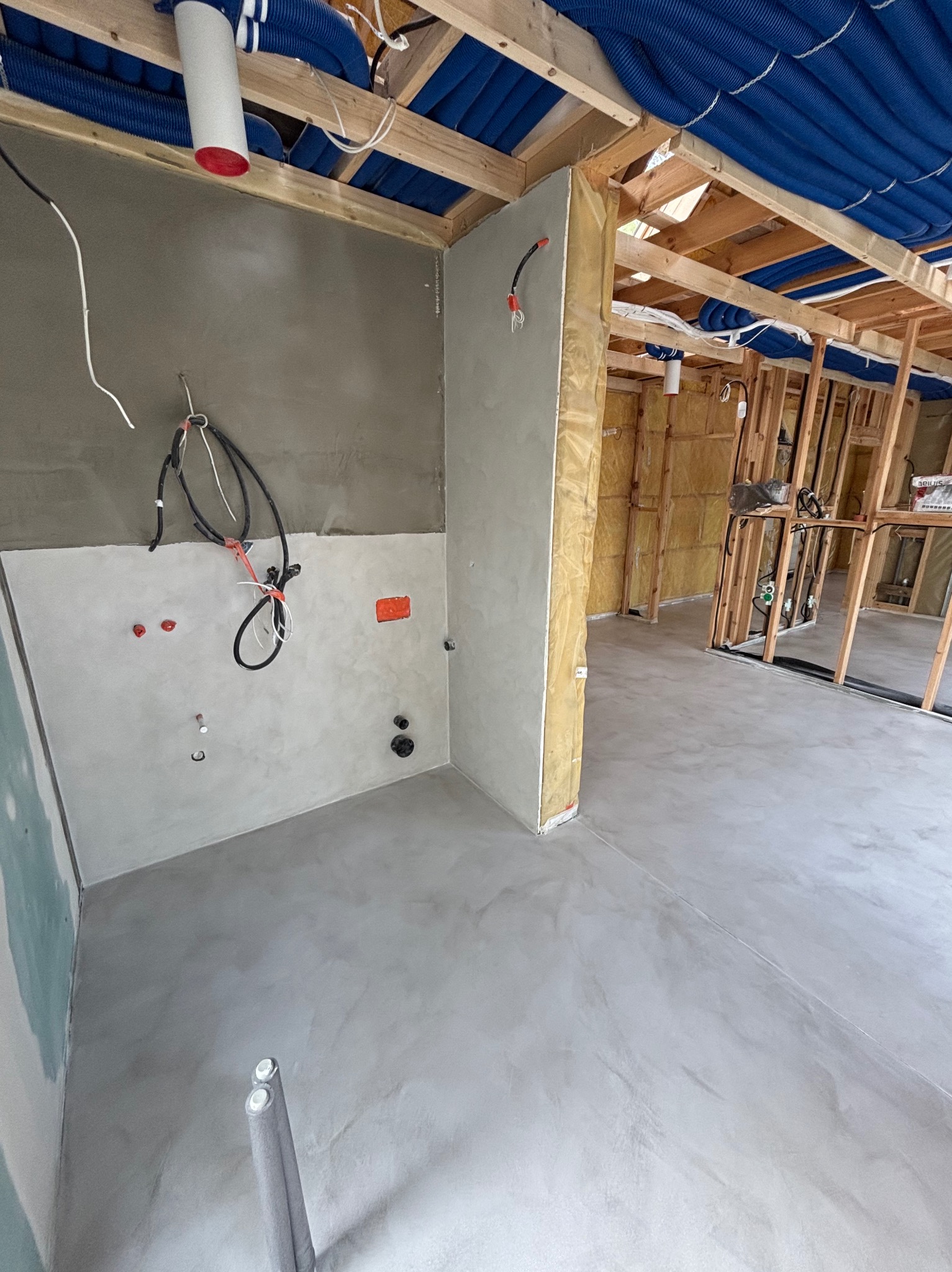 Szara, gładka posadzka w surowym wnętrzu. Instalacje pod sufitem i ściany w trakcie wykończenia. Widoczne przewody i rury.