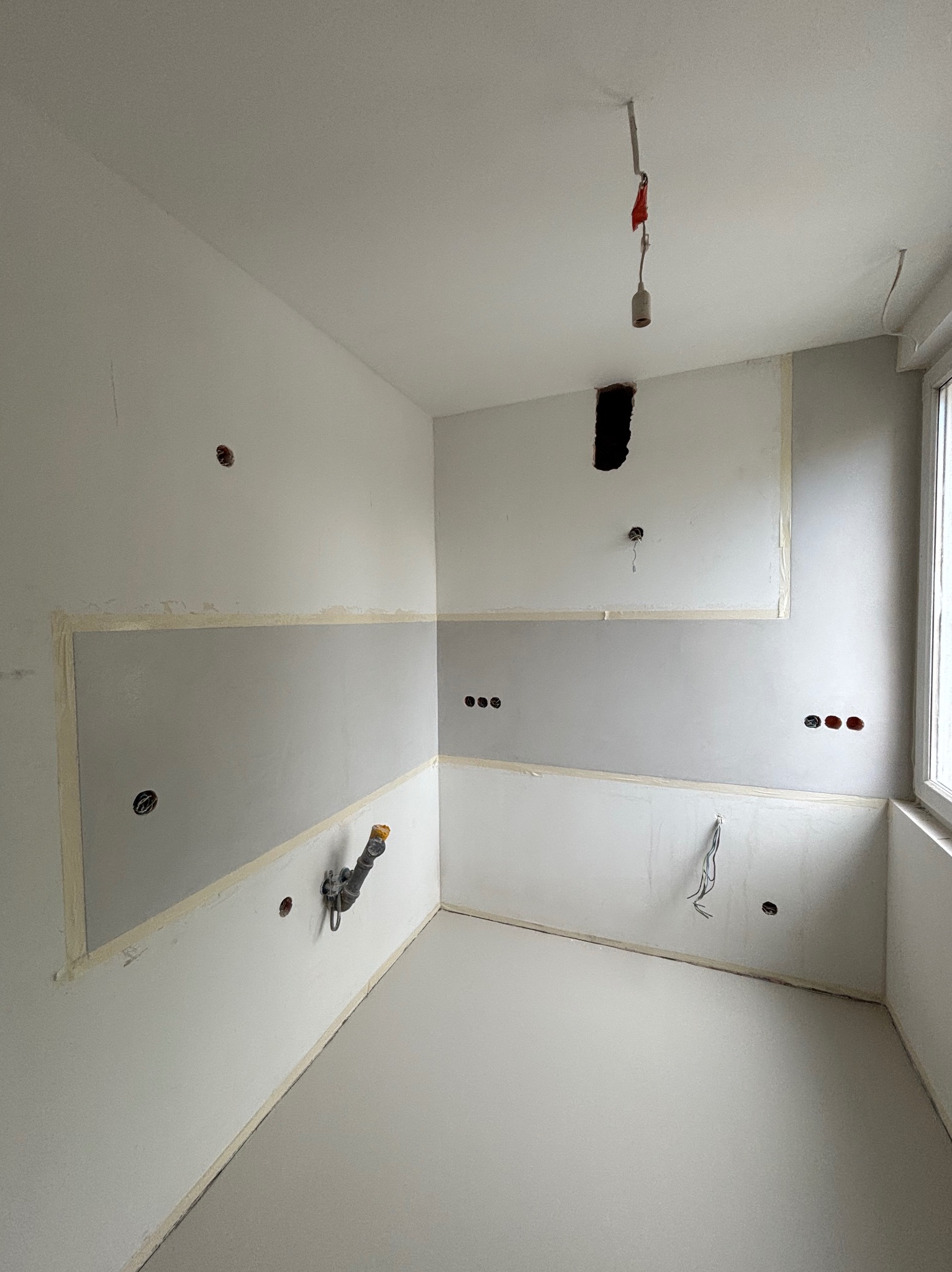 Wnętrze w trakcie prac wykończeniowych: szara ściana zabezpieczona taśmą, świeża posadzka, instalacje elektryczne i hydrauliczne, surowa żarówka.