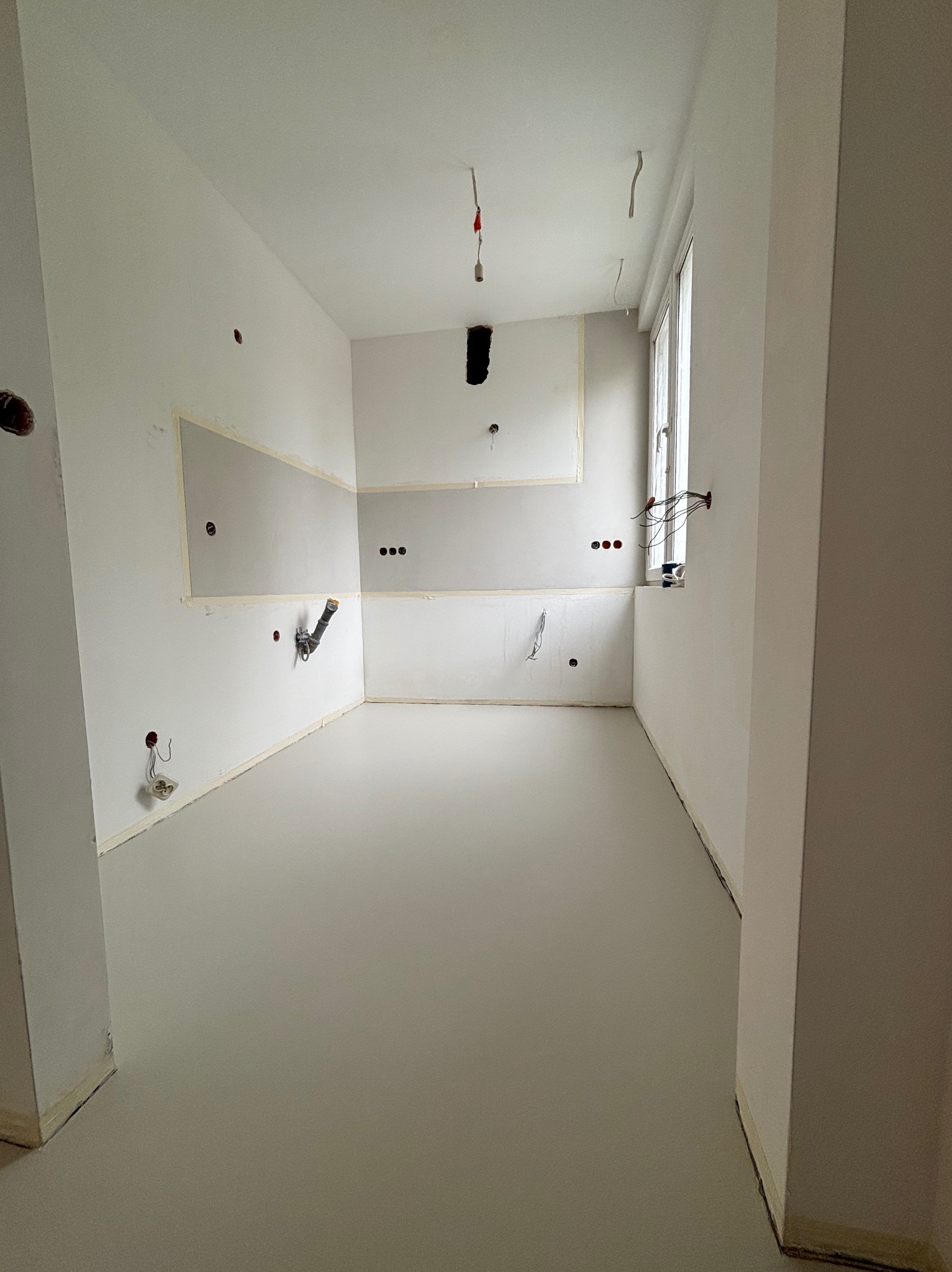 Jasnoszara, gładka posadzka w pustym pomieszczeniu z przygotowanymi instalacjami na ścianach i oknem w tle. Widoczne taśmy malarskie.