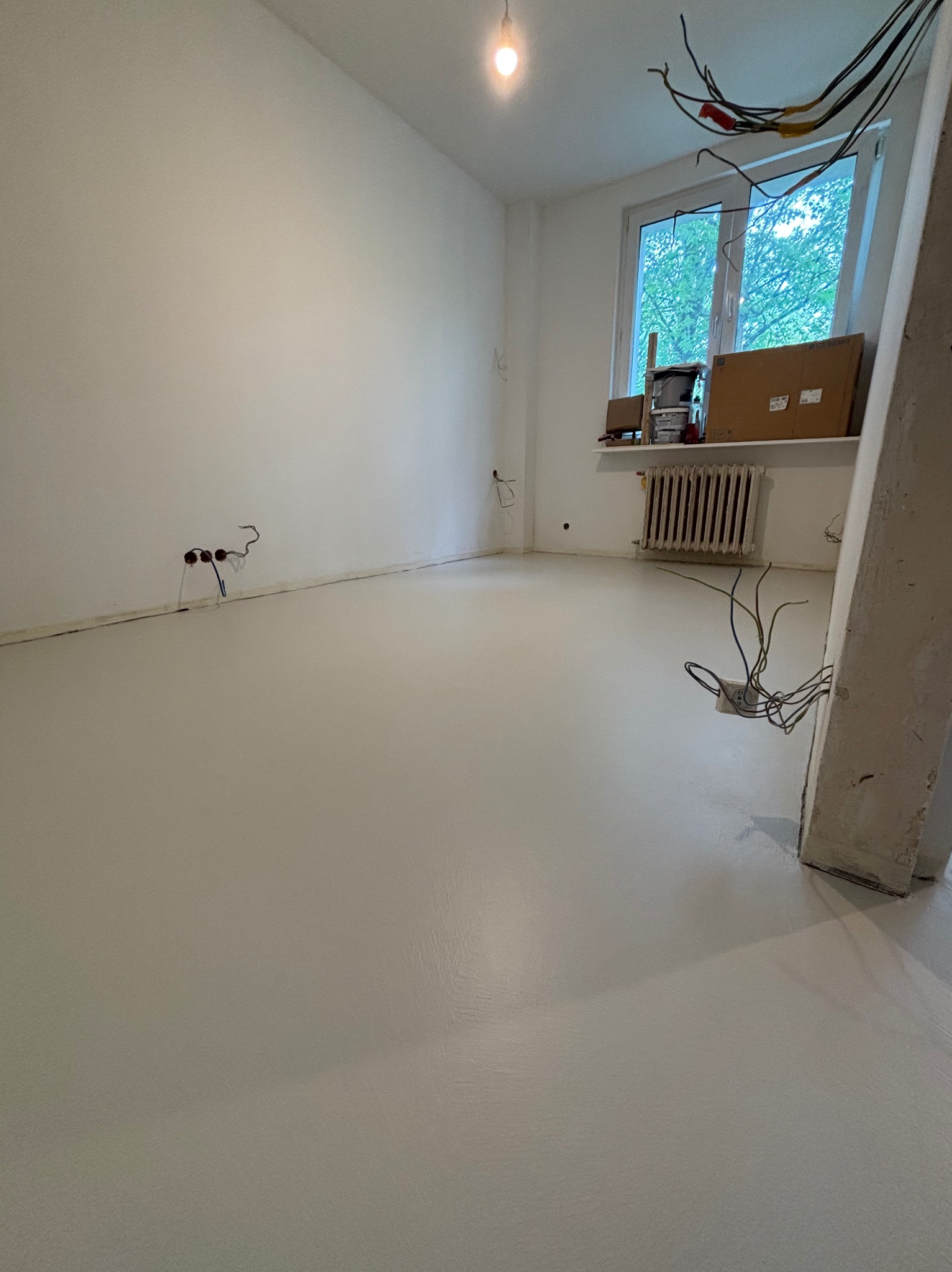Jasnoszara, gładka posadzka w pustym pokoju z grzejnikiem pod oknem. Widoczne wystające kable ze ściany i sufitu. Świeżo wylana posadzka w trakcie prac wykończeniowych.