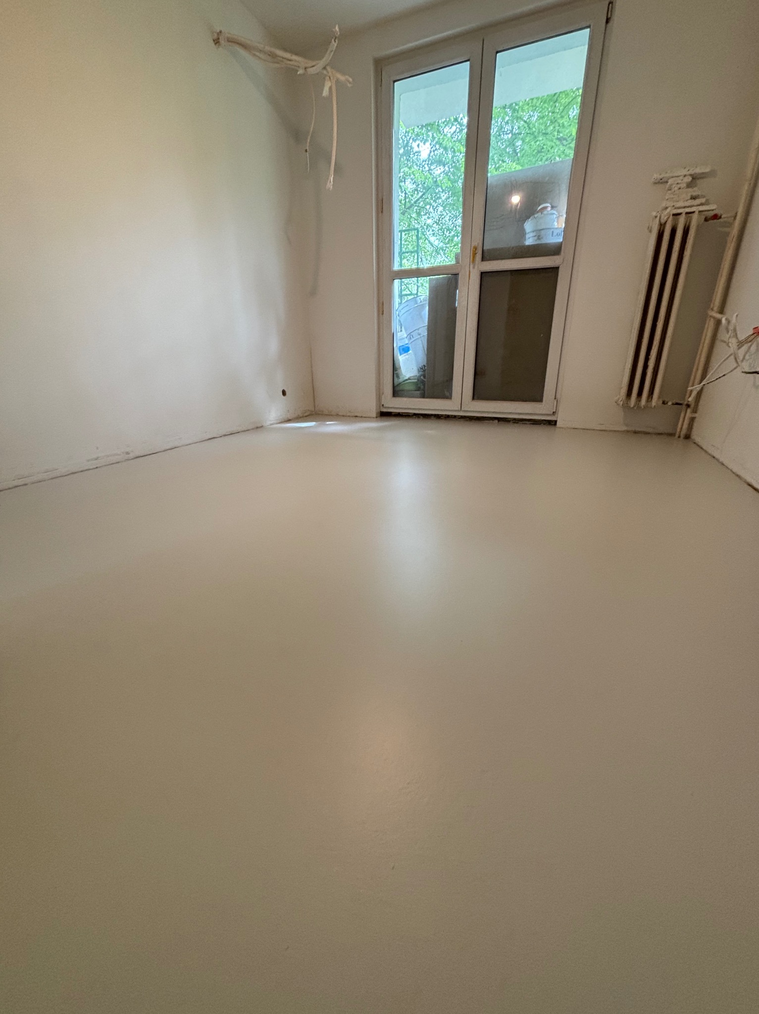 Jasna, gładka posadzka w pokoju z białymi ścianami i oknem balkonowym. Grzejnik żeliwny przy ścianie. Widoczne przewody elektryczne wystające ze ściany.