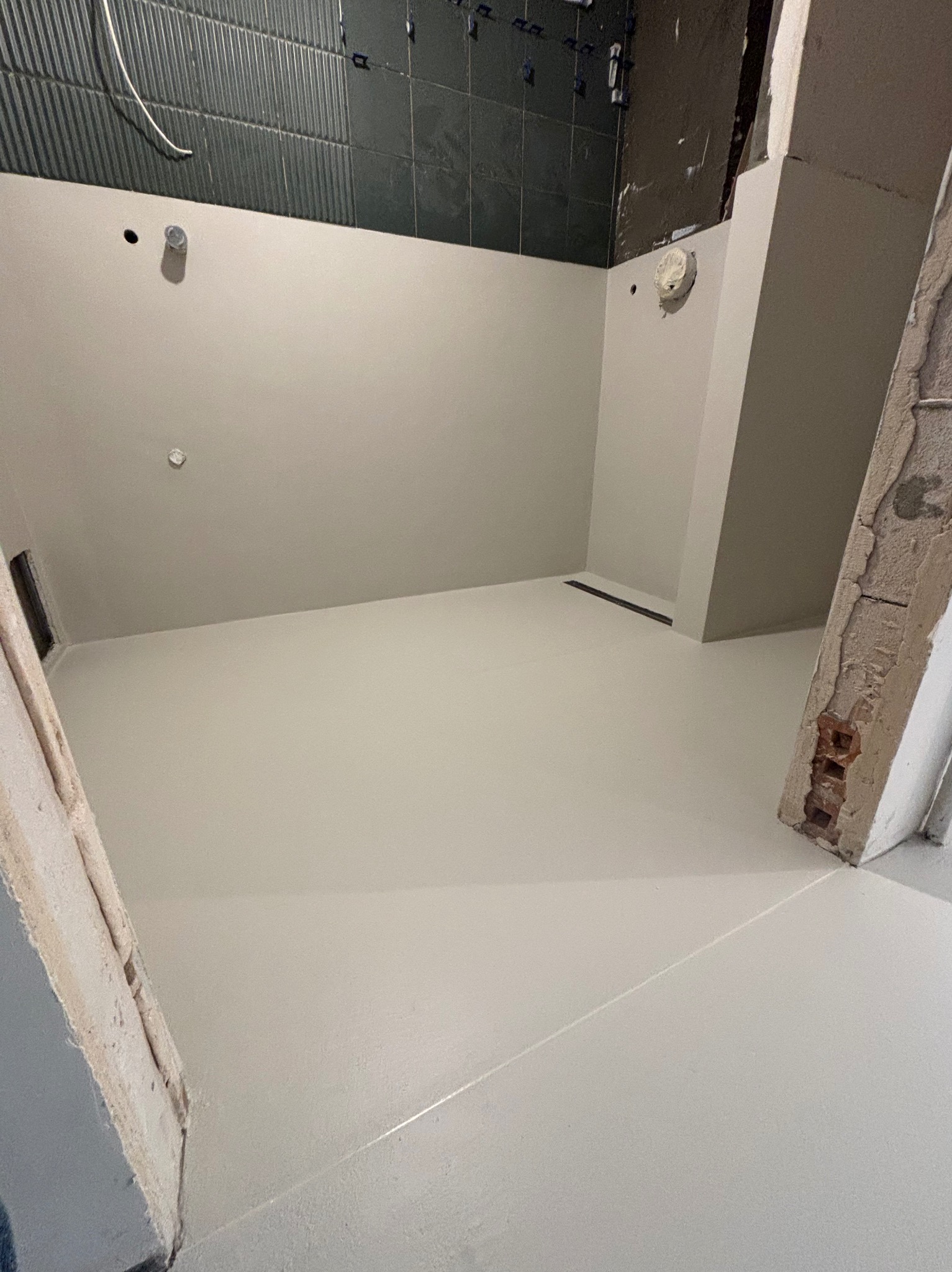 Jasna, gładka posadzka w łazience z ciemnymi płytkami na ścianie i odpływem liniowym. Widoczne surowe wykończenia ścian.