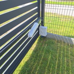 KEY DOOR Automatyka Bramowa - Szara, pozioma brama lamelowa z zamontowanym napędem RAY2524EK i skrzynką sterującą na słupku. Widok na trawnik i ogrodzenie z siatki.