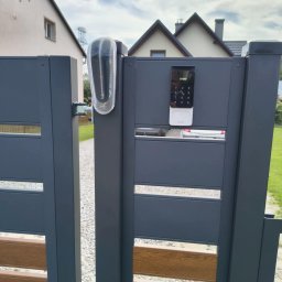KEY DOOR Automatyka Bramowa - Nowoczesna, grafitowa brama z drewnopodobnymi panelami, wyposażona w minimalistyczny wideodomofon z klawiaturą numeryczną i lampę ostrzegawczą.