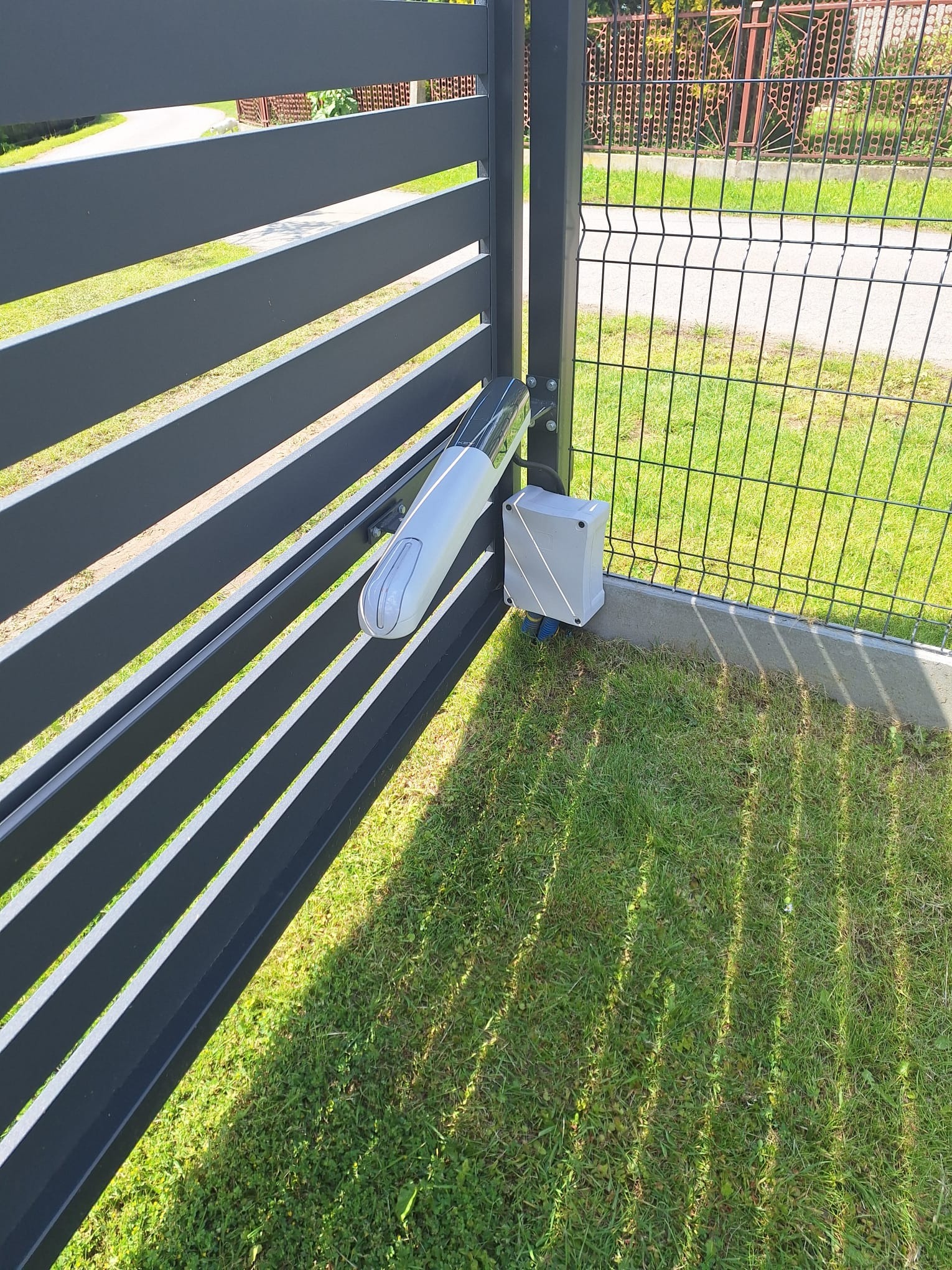 Szara, pozioma brama lamelowa z zamontowanym napędem RAY2524EK i skrzynką sterującą na słupku. Widok na trawnik i ogrodzenie z siatki.