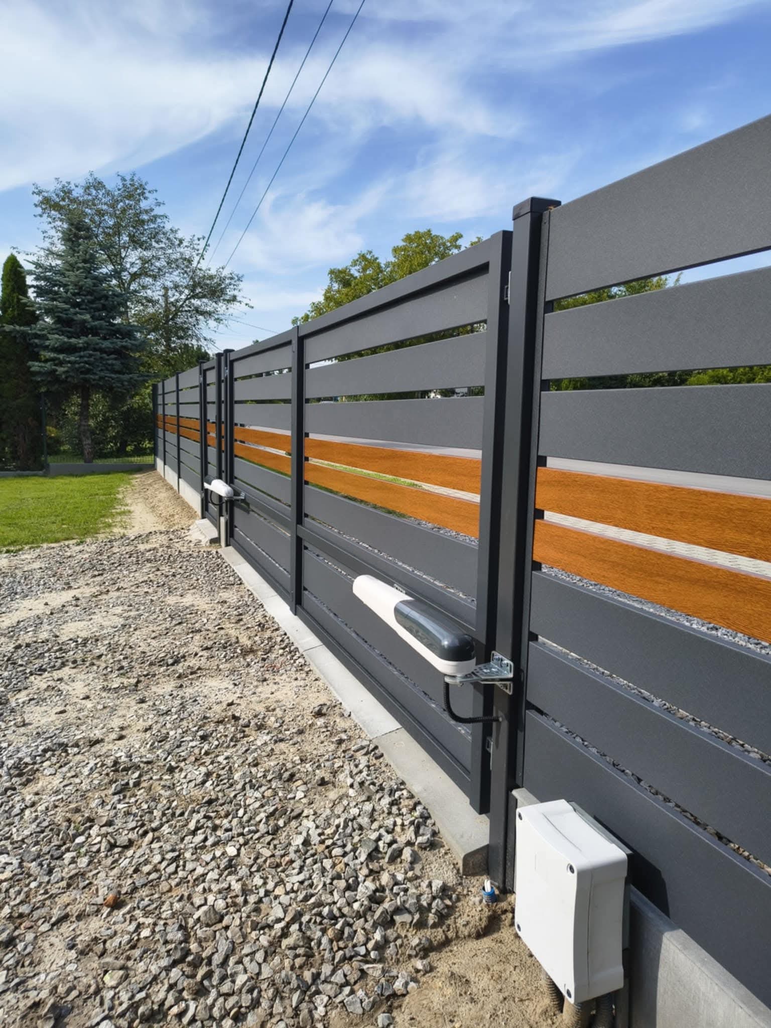 Nowoczesna brama wjazdowa z automatyką RAY2524EK, szara z drewnianymi akcentami, widoczny napęd i skrzynka sterownicza na tle żwirowego podjazdu i zieleni.