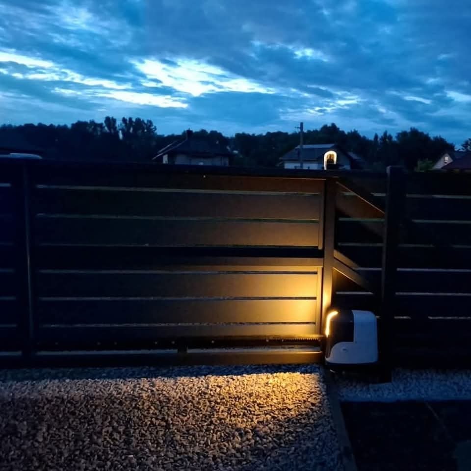 Nowoczesna, przesuwna brama wjazdowa z automatyką, oświetlona od dołu, na tle wieczornego nieba. Widoczny napęd bramy i żwirowe podłoże.