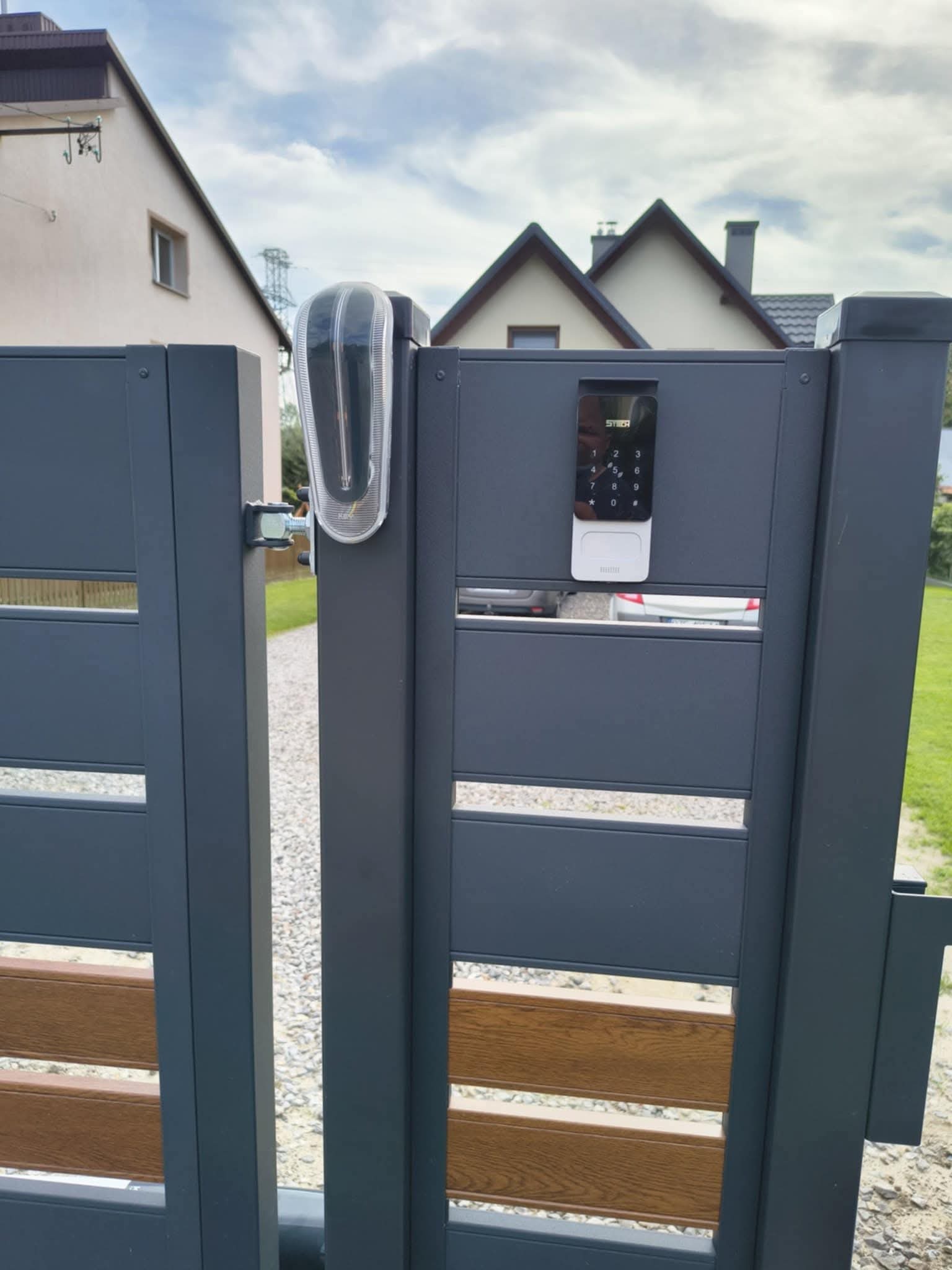 Nowoczesna, grafitowa brama z drewnopodobnymi panelami, wyposażona w minimalistyczny wideodomofon z klawiaturą numeryczną i lampę ostrzegawczą.