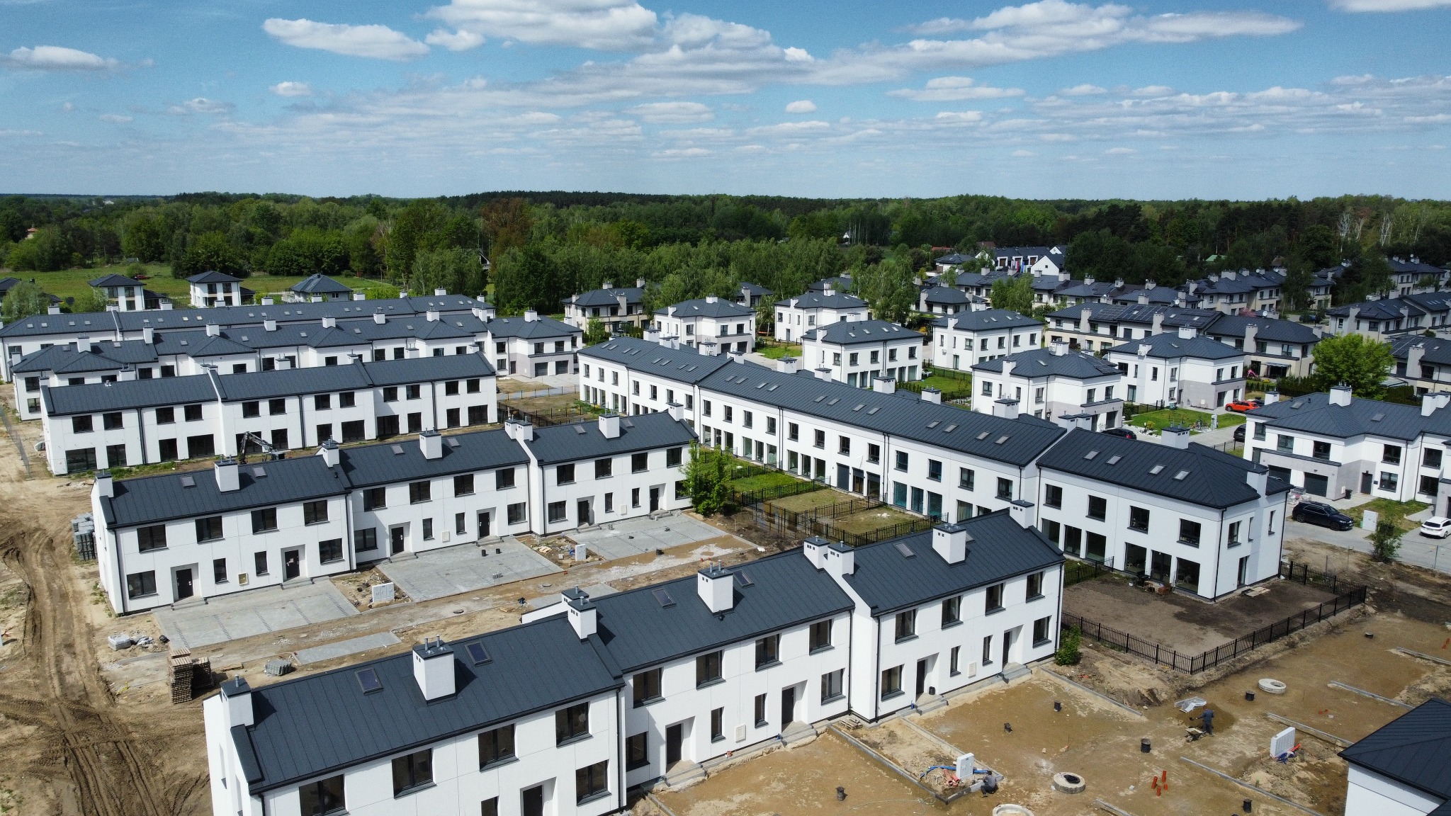 Panoramiczny widok z lotu ptaka na osiedle nowych, białych domów z ciemnymi dachami, częściowo w trakcie budowy, otoczonych zielenią i błękitnym niebem.