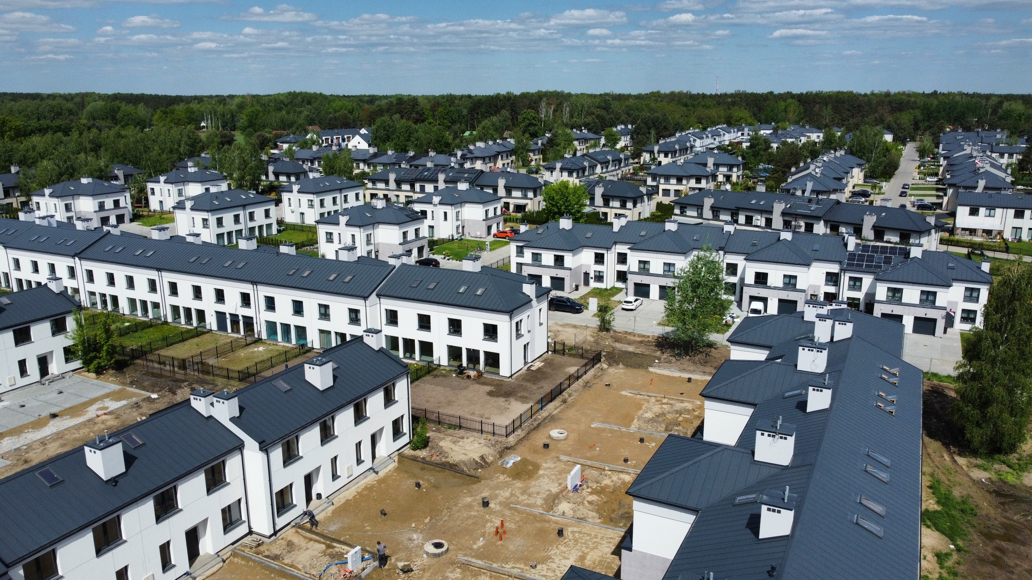 Osiedle nowych, dwukondygnacyjnych domów w zabudowie szeregowej z ciemnymi dachami, widziane z lotu ptaka. Część działek w trakcie zagospodarowania.