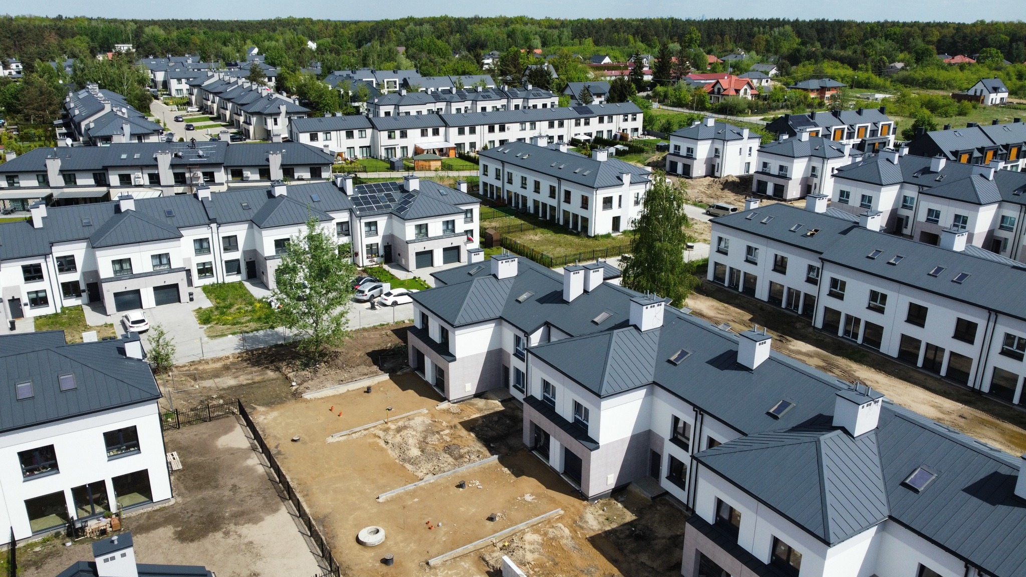 Osiedle nowoczesnych domów z szarymi dachami, widok z góry. Część budynków w stanie surowym, inne wykończone z garażami i podjazdami. W tle zieleń drzew.