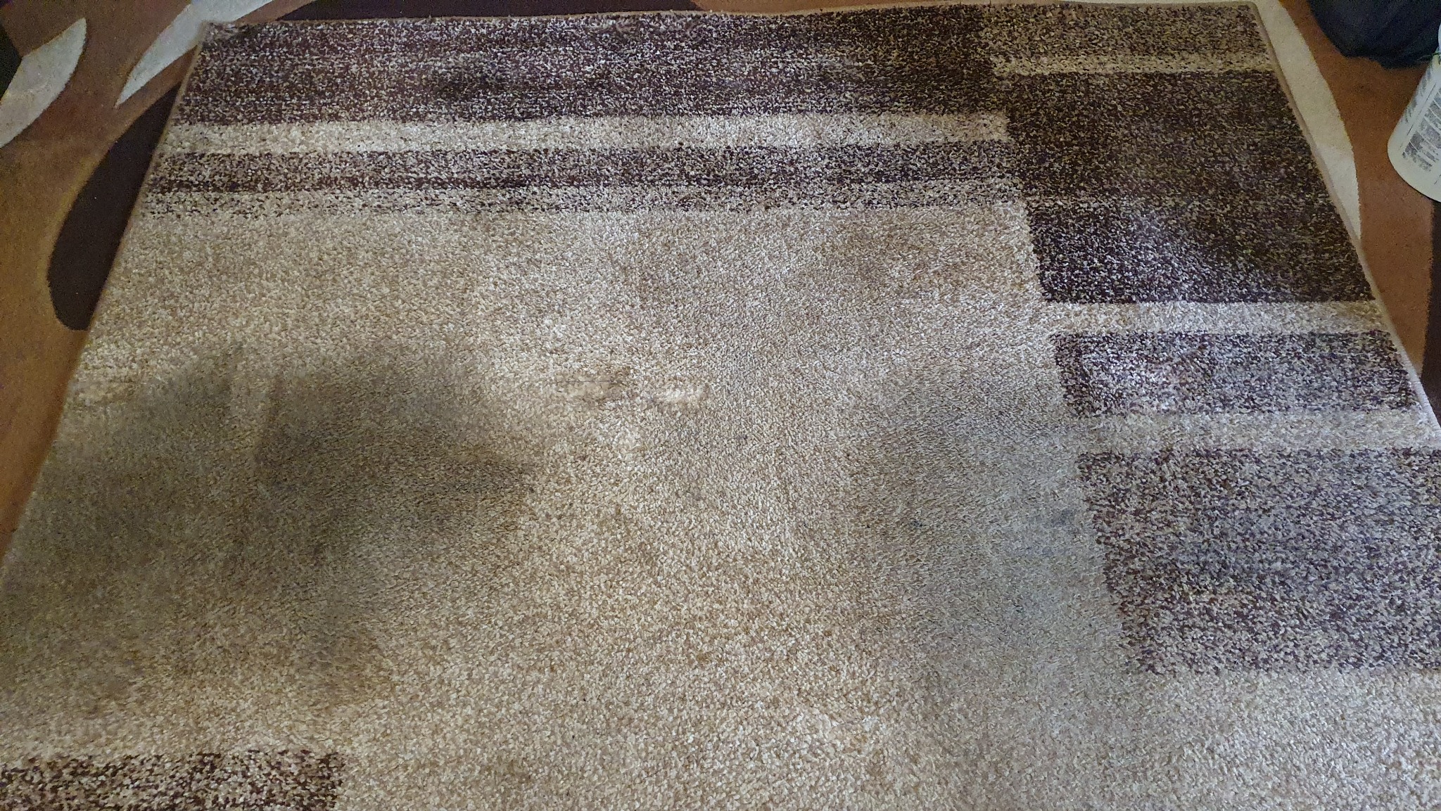 Beżowo-brązowy dywan z geometrycznym wzorem, widoczne zabrudzenia. Zdjęcie dywanu przed czyszczeniem, z bliska, w dziennym świetle.