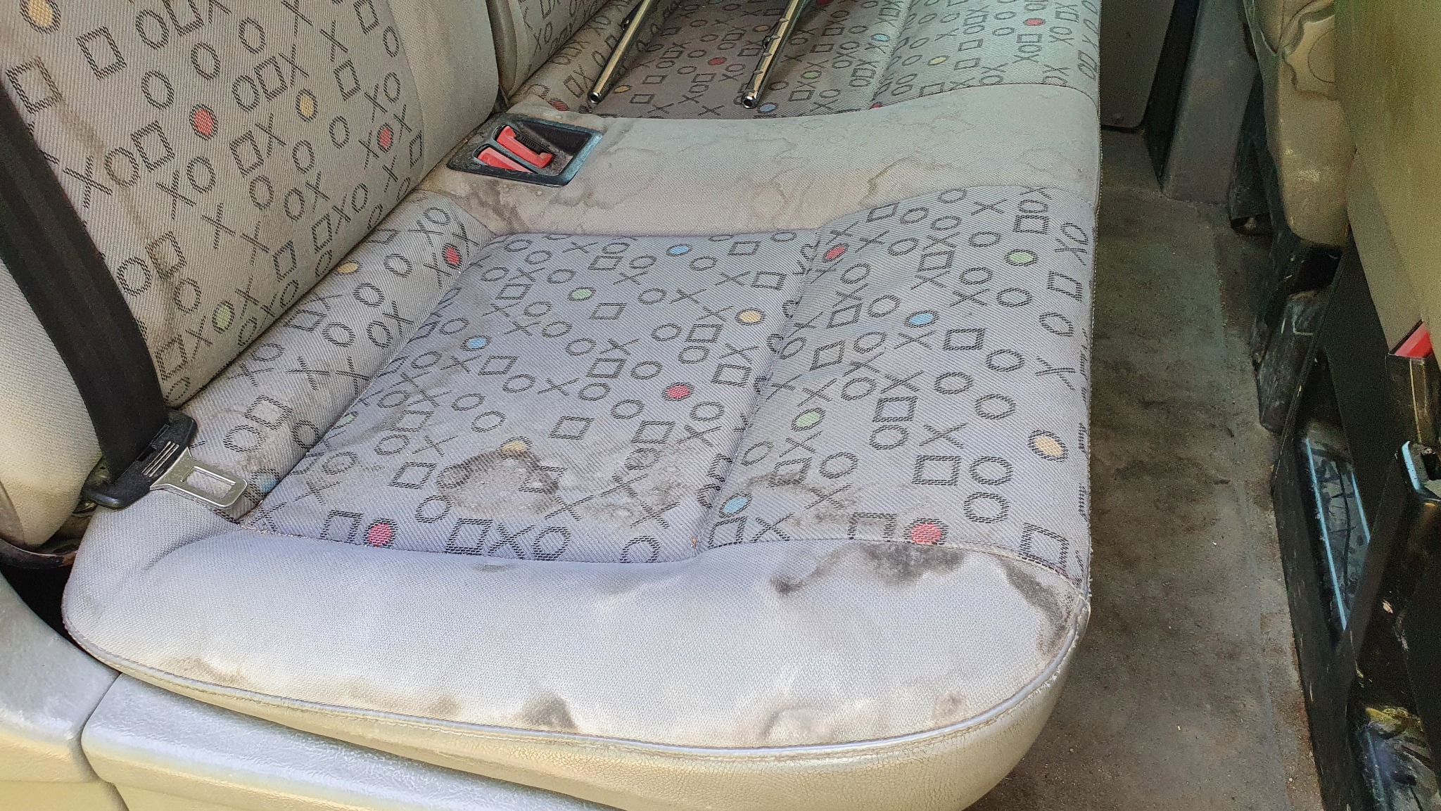 Silnie zabrudzone, szare siedzenie samochodowe z wzorem kółek, krzyżyków i kwadratów przed czyszczeniem. Widoczne plamy i zabrudzenia na tapicerce.