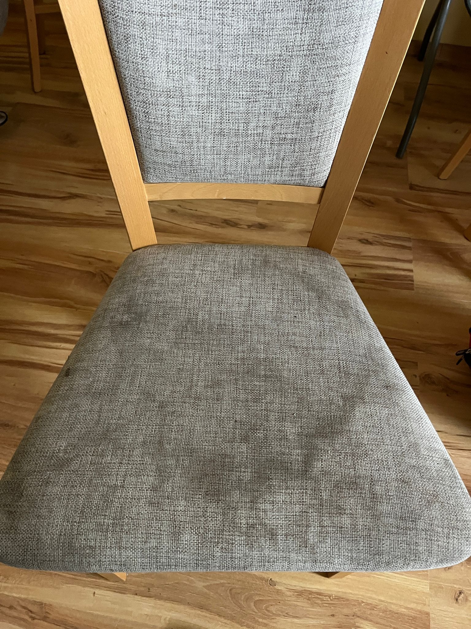 Krzesło z szarą tapicerką, widoczne plamy na siedzisku. Ujęcie z góry, drewniana rama krzesła i parkiet w tle. Zdjęcie oddaje stan tapicerki przed czyszczeniem.